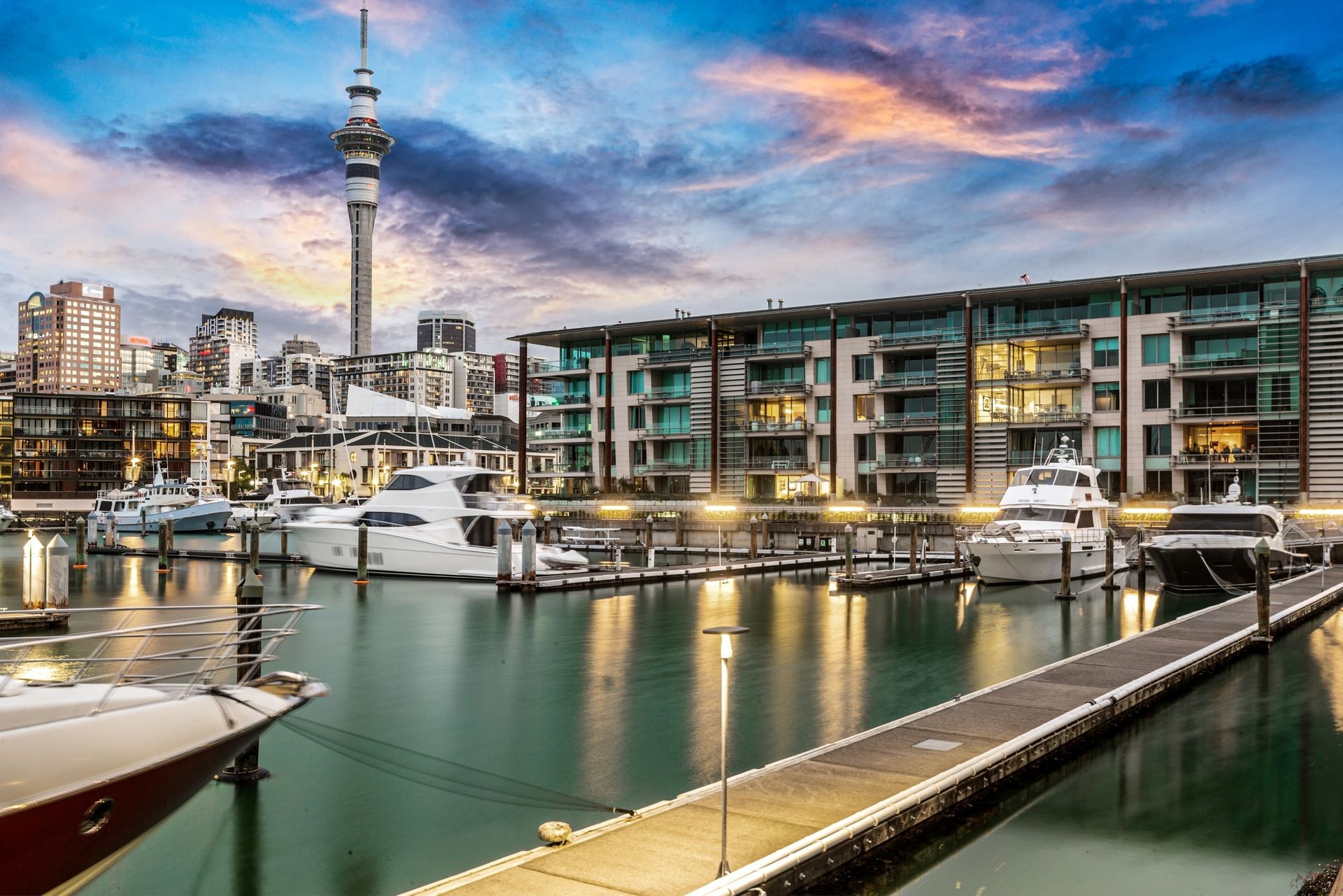 Auckland harbor with boats, buildings, and Sky Tower at sunset.