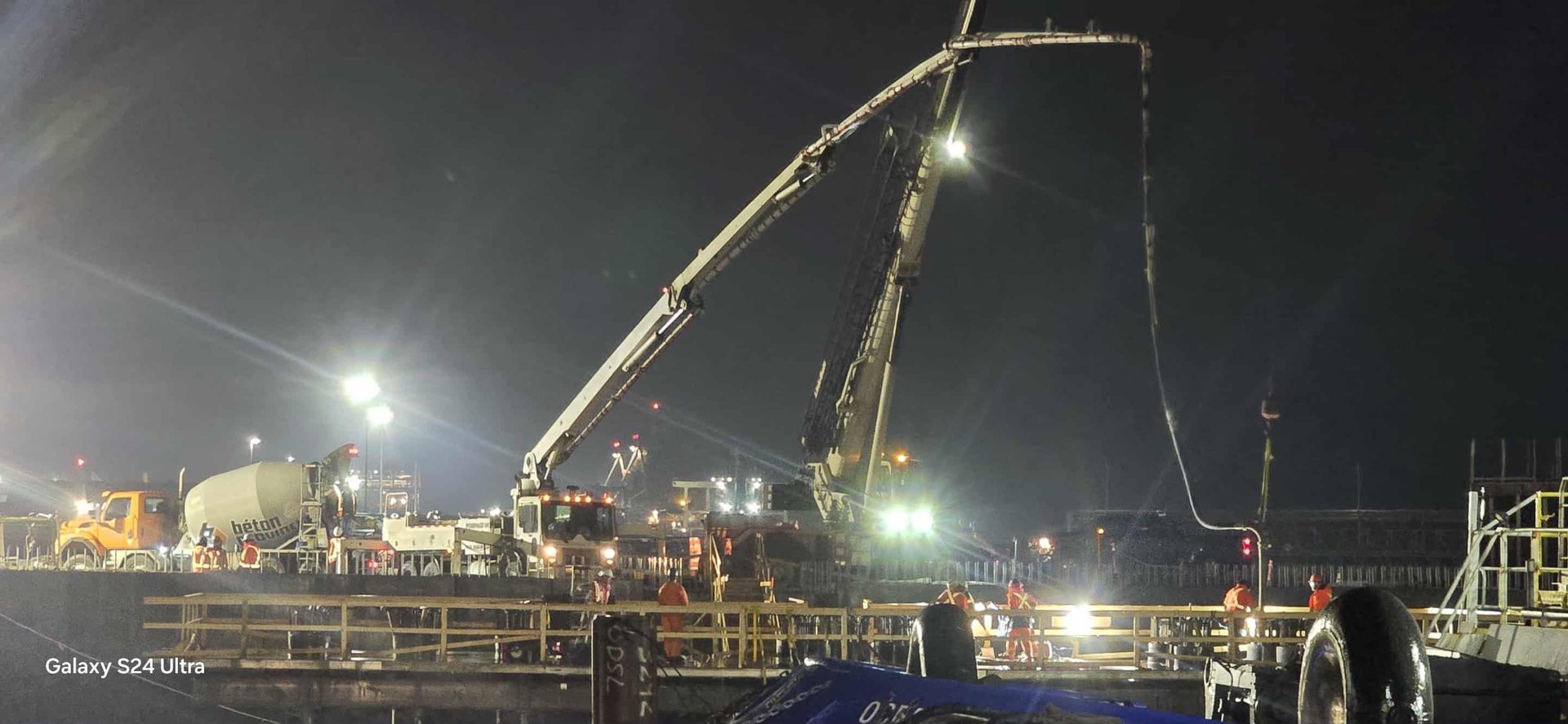 Chantier de construction de nuit : Pompe à béton en action, camions et ouvriers éclairés par des projecteurs puissants.