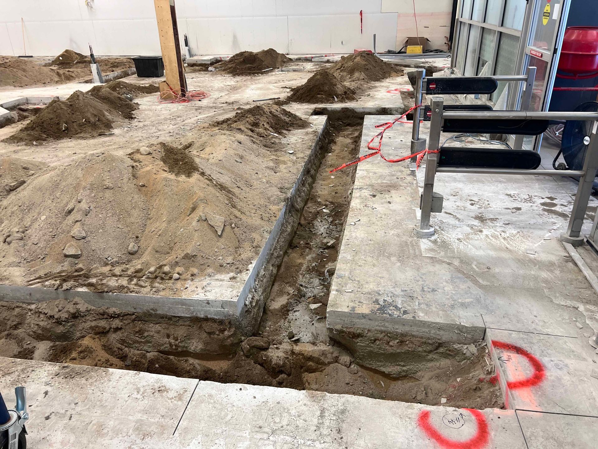 Chantier de construction avec une longue tranchée creusée dans un sol en béton, des tas de terre à proximité.