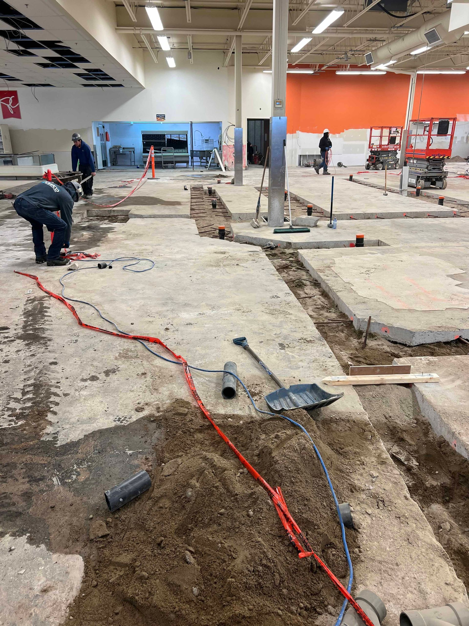 Travaux de construction dans un grand magasin : ouvriers portant des casques de chantier, sols en béton apparent, tranchées, outils, câbles rouges et bleus.