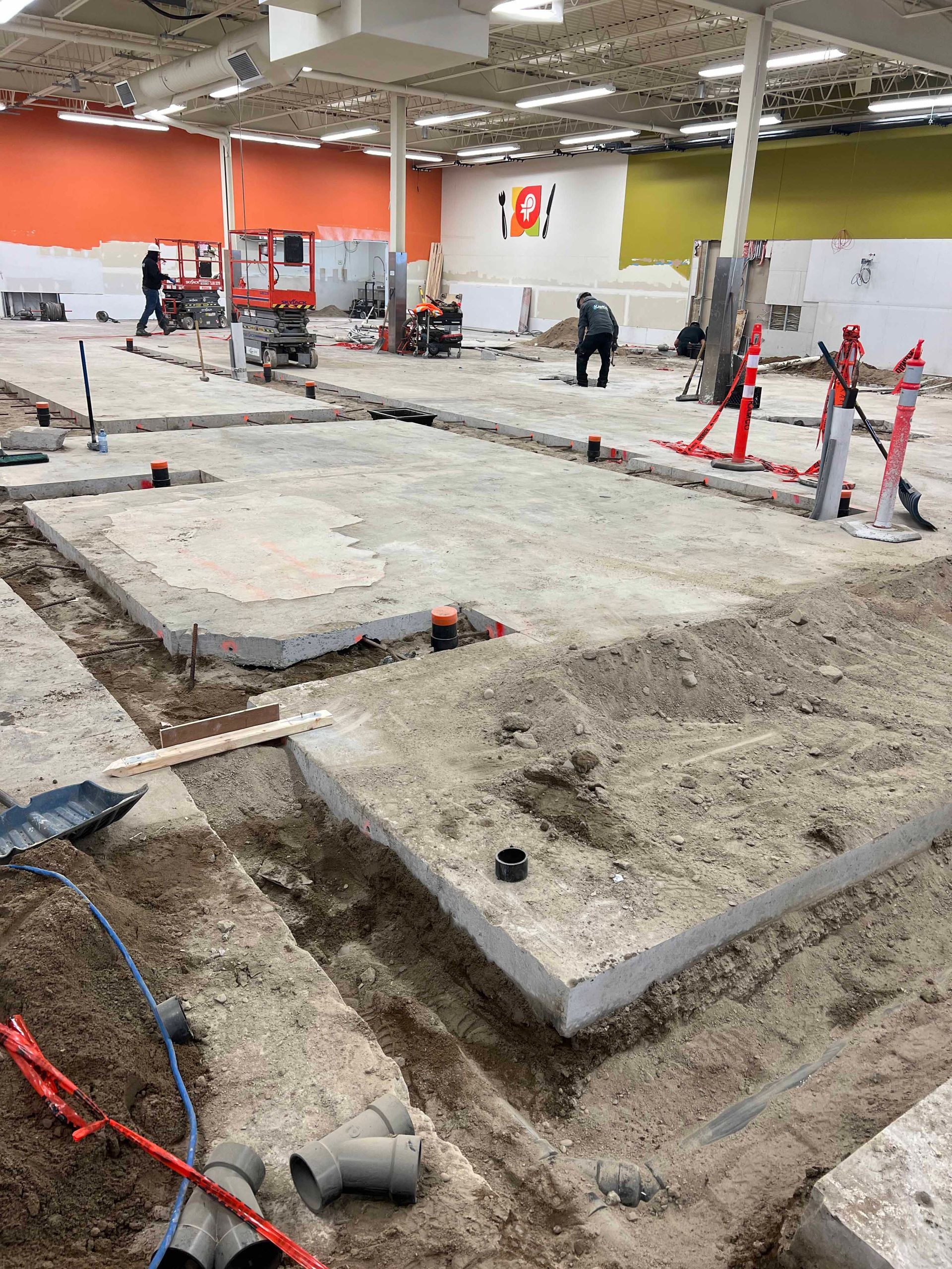 Chantier : Ouvriers à l'intérieur d'un grand bâtiment aux dalles de béton et aux canalisations apparentes. Murs orange et jaunes.