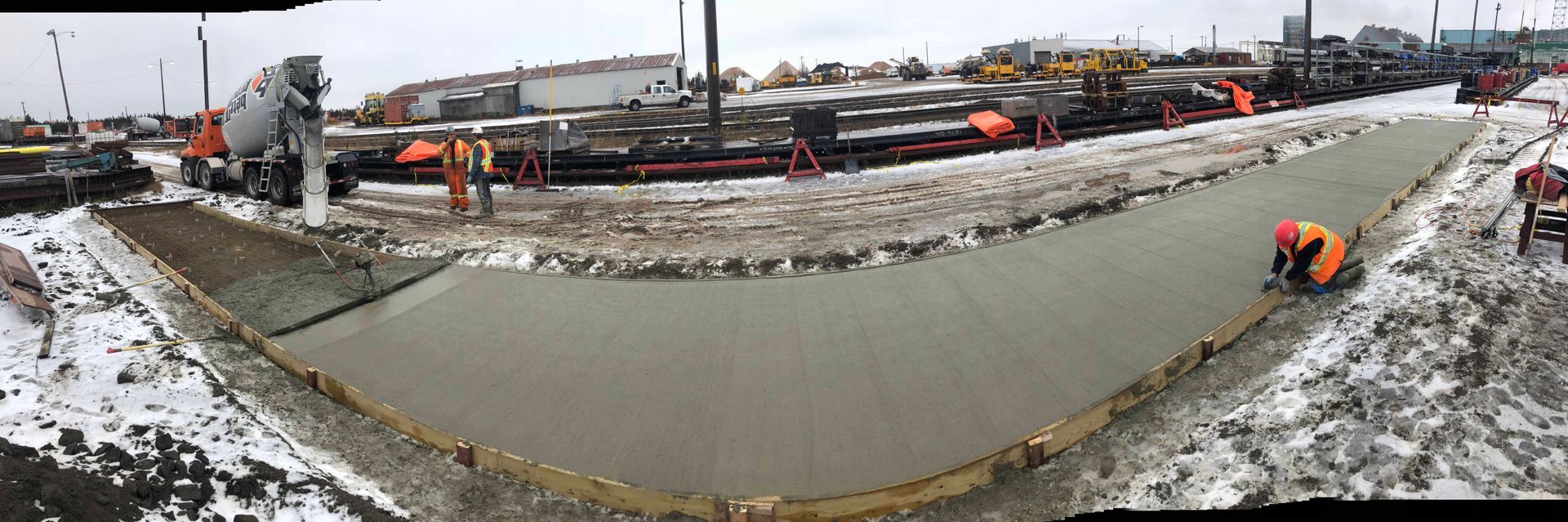 Chantier de construction avec des ouvriers coulant du béton pour une route, entouré de neige et d'engins de chantier.