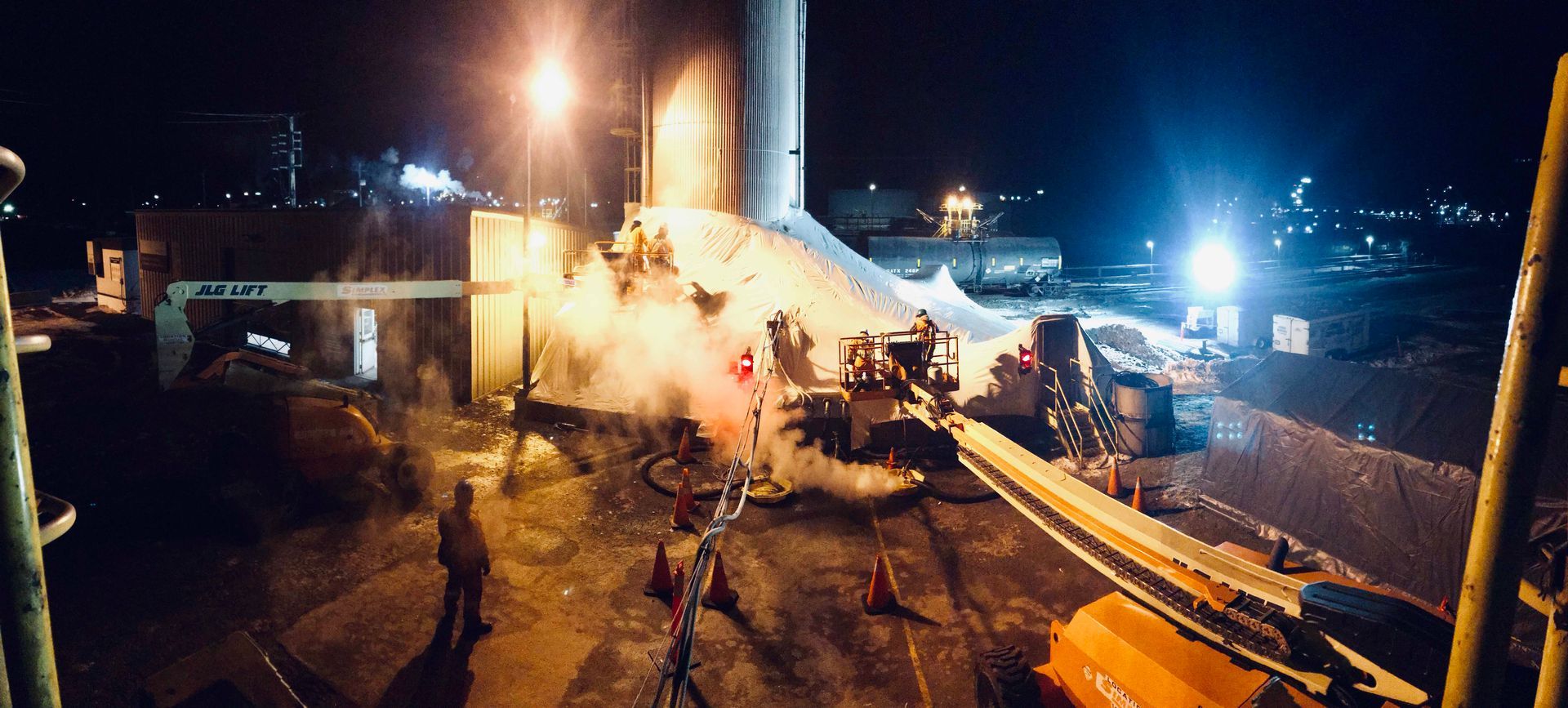Scène industrielle nocturne avec des ouvriers près d'une grande structure. Lumières vives, vapeur et machines sont visibles.
