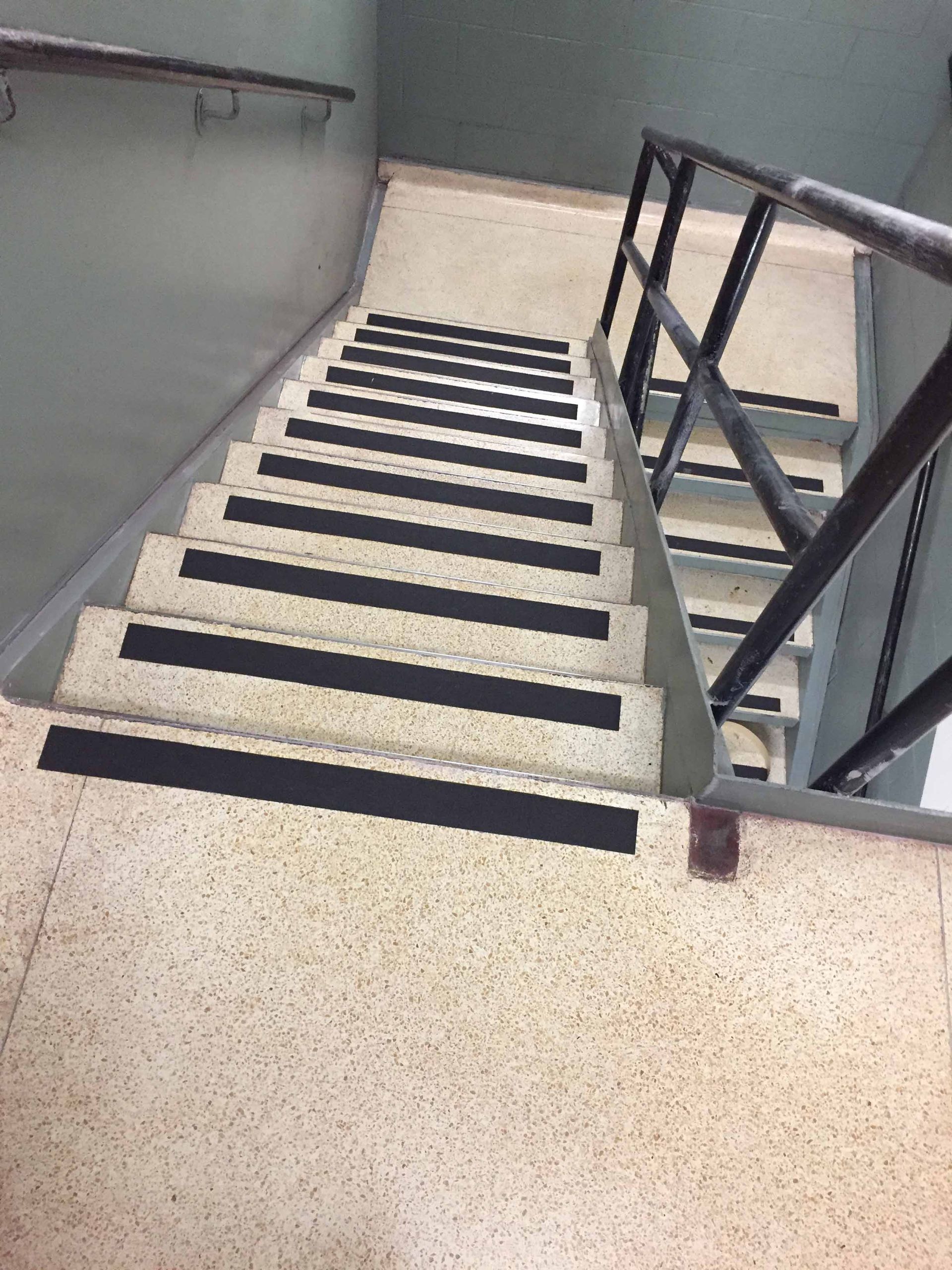 Escalier avec bandes antidérapantes noires sur chaque marche, rampes métalliques et sol et murs en terrazzo de couleur neutre.