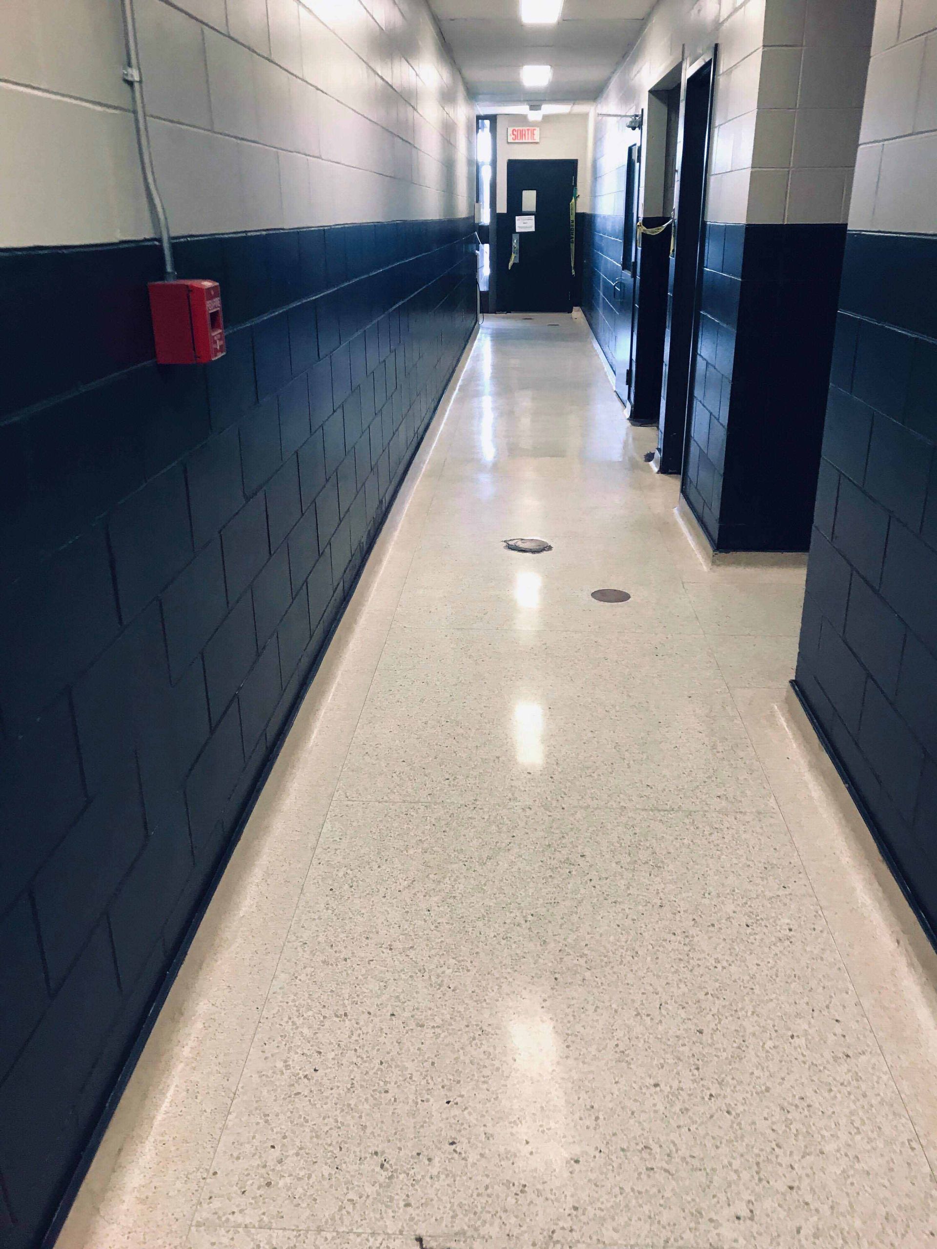 Long couloir aux murs blancs et bleu marine, sol moucheté, alarme incendie et portes.