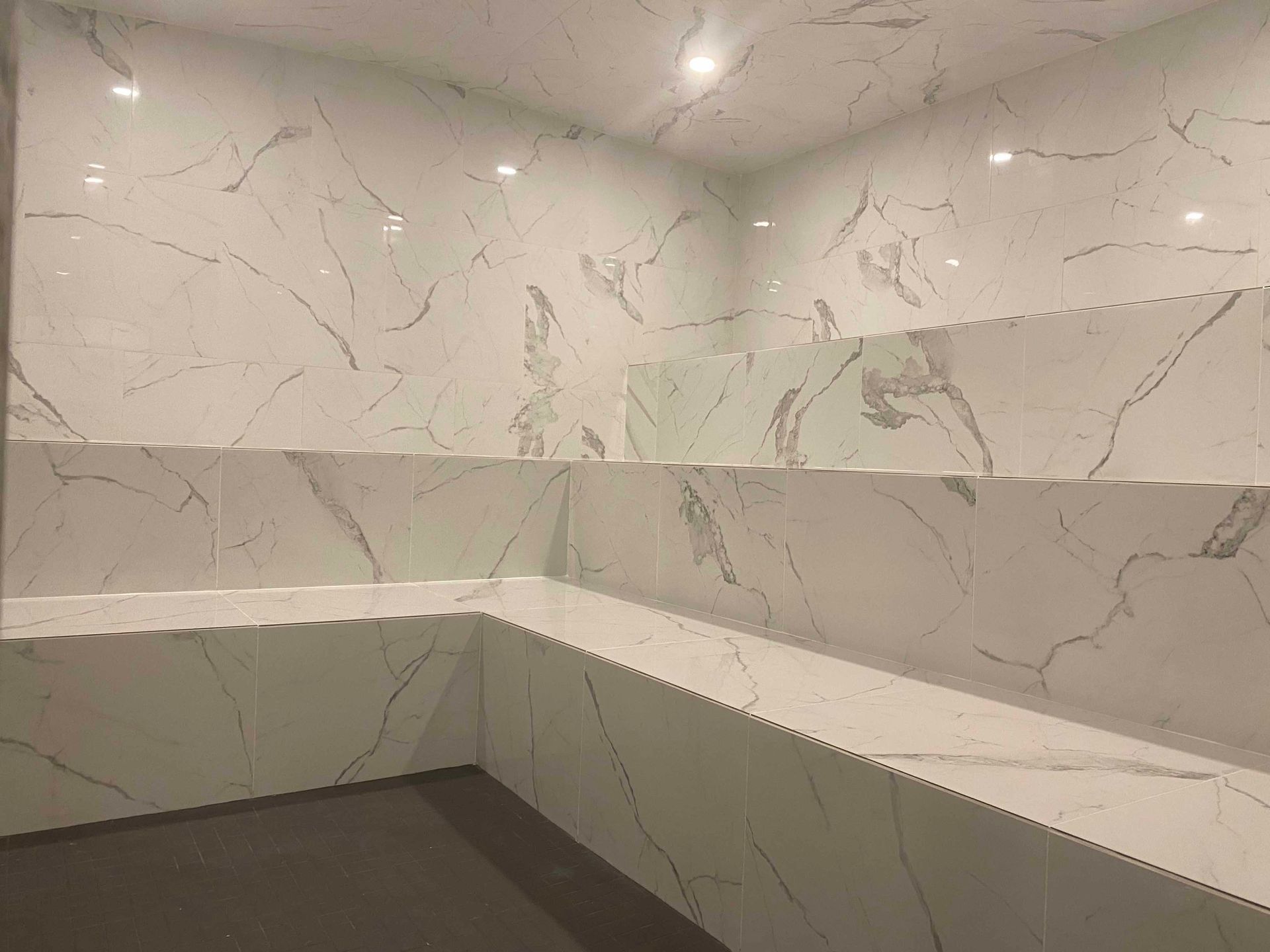 Douche carrelée en marbre blanc avec étagères intégrées. Des veines grises parcourent les carreaux. Éclairage encastré.