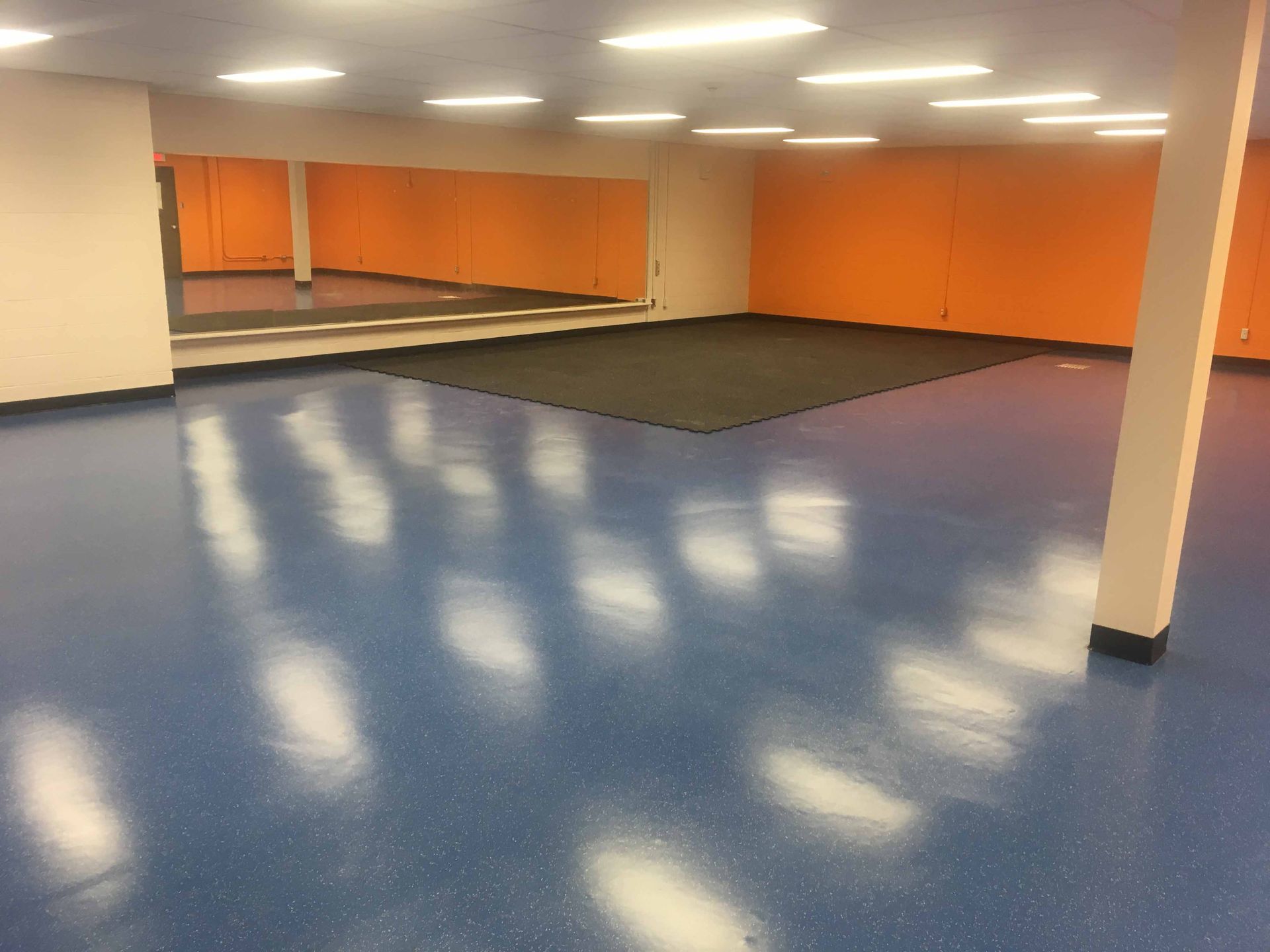 Studio de danse avec sol bleu, murs orange, miroirs et tapis noir.