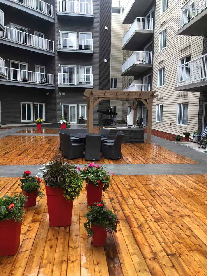 Terrasse extérieure avec plancher en bois, coin salon sous une pergola et pots de fleurs rouges.
