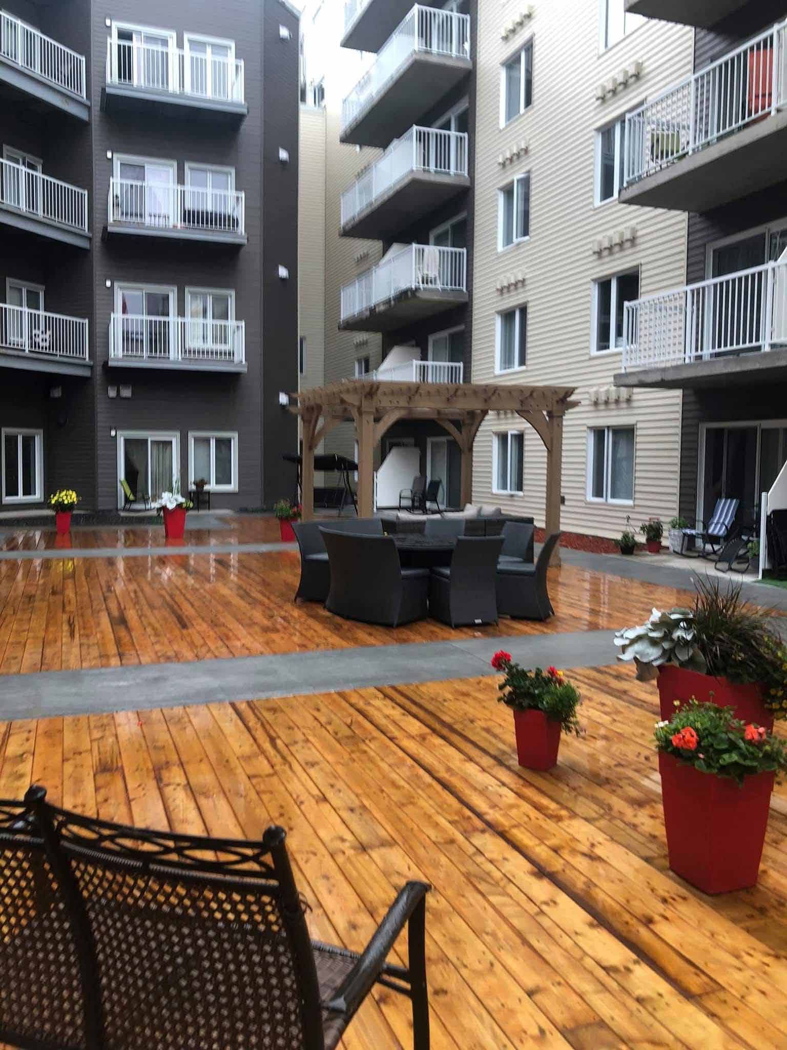 Cour intérieure avec terrasse en bois, mobilier de jardin, pergola et immeubles d'appartements en arrière-plan. Des pots de fleurs rouges agrémentent l'espace.