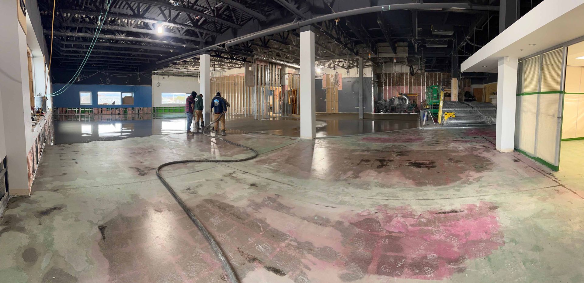 Vue intérieure d'un espace industriel inondé, probablement en construction ; des ouvriers au centre avec un tuyau d'arrosage, plafond sombre et sol mouillé.