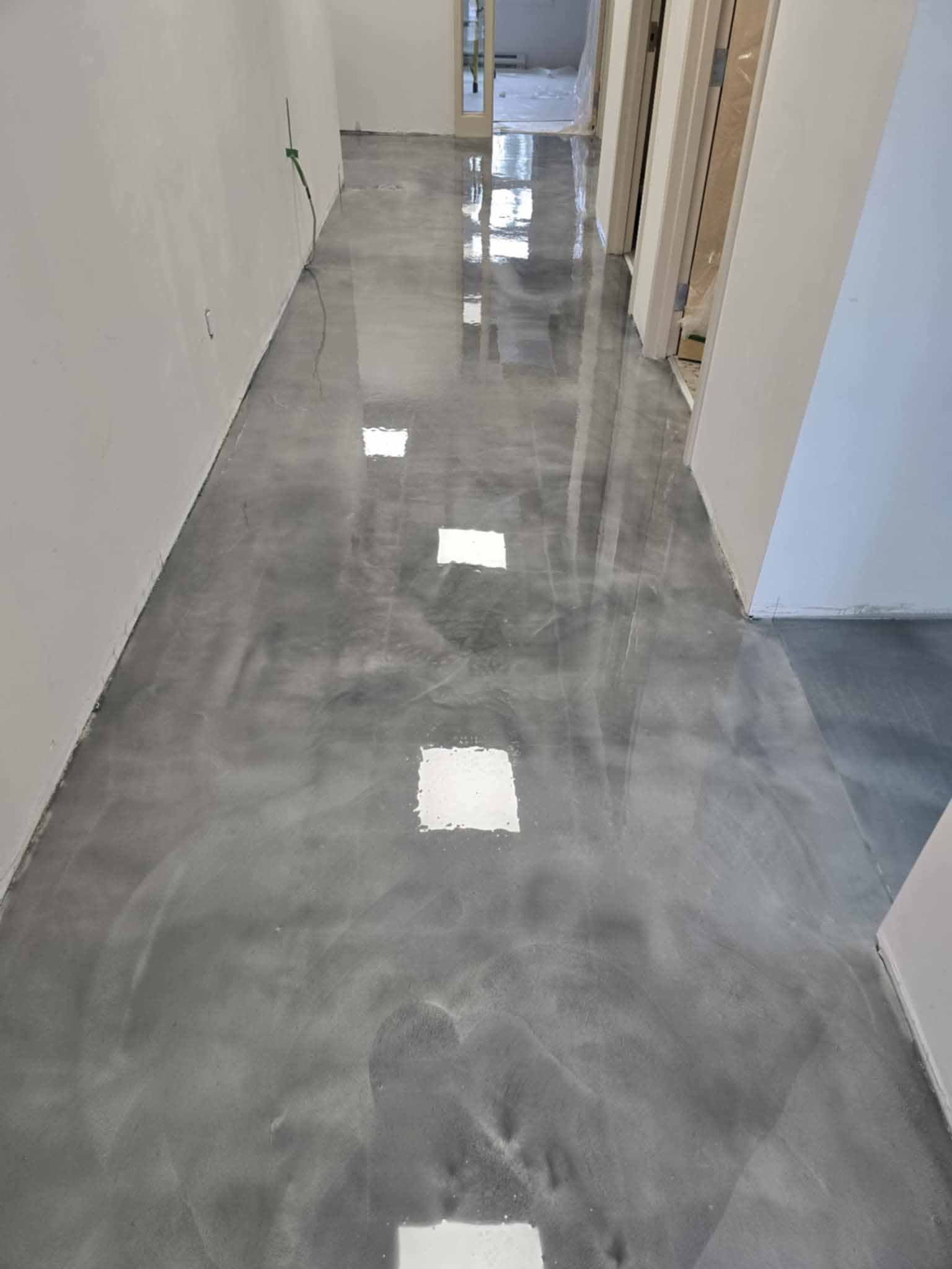 Un long couloir au sol en époxy gris brillant. Les murs sont blancs et bruts, avec des portes.