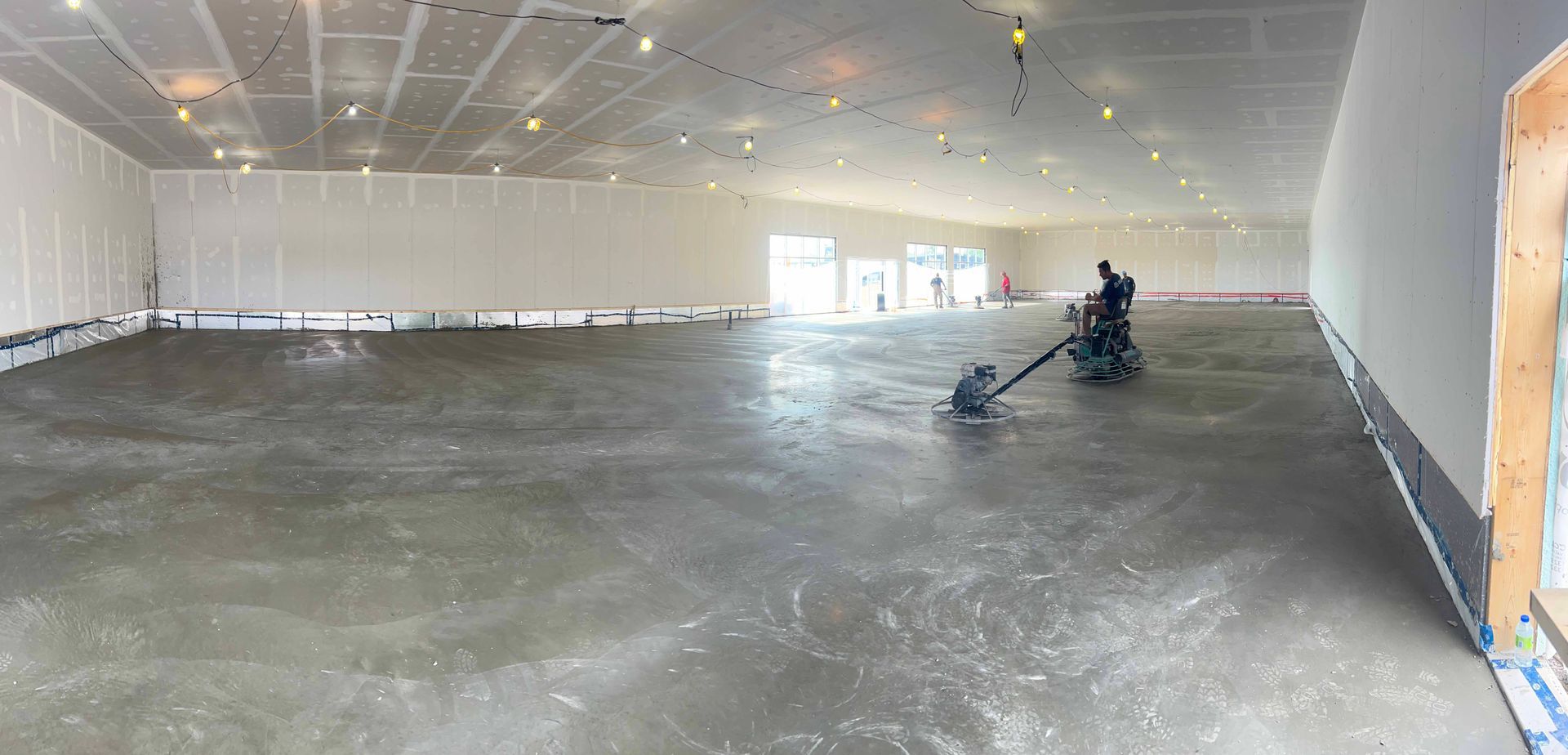 Une personne lisse un sol en béton dans un grand espace de construction ouvert. Les murs sont blancs et le plafond est équipé de spots encastrés.