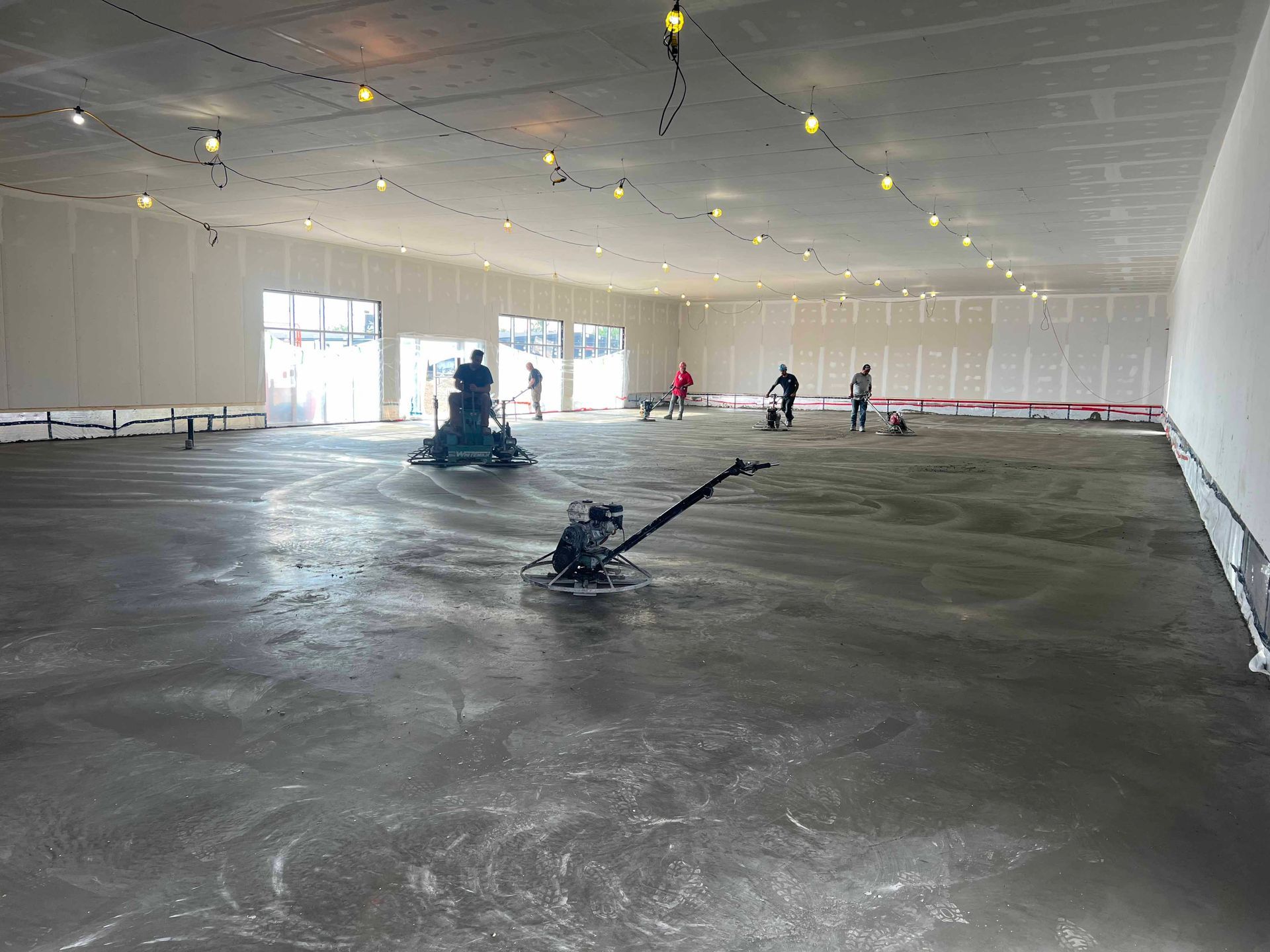 Des ouvriers lissent un sol en béton dans un grand espace commercial ouvert, avec un plafond apparent et des guirlandes lumineuses.