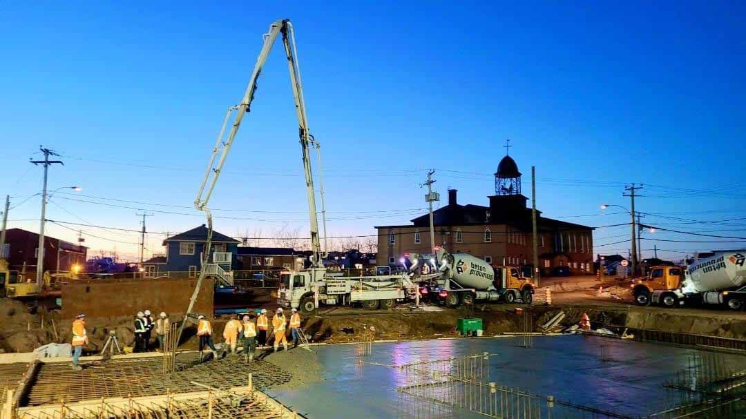 Chantier au crépuscule : coulage de béton avec camion-pompe, ouvriers et un bâtiment historique en briques en arrière-plan.