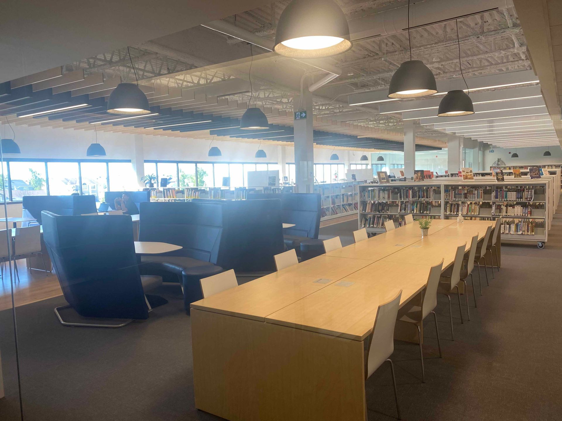 Intérieur d'une bibliothèque avec une longue table, des chaises, des étagères et divers espaces de détente. Éclairage assuré par plusieurs lampes suspendues.