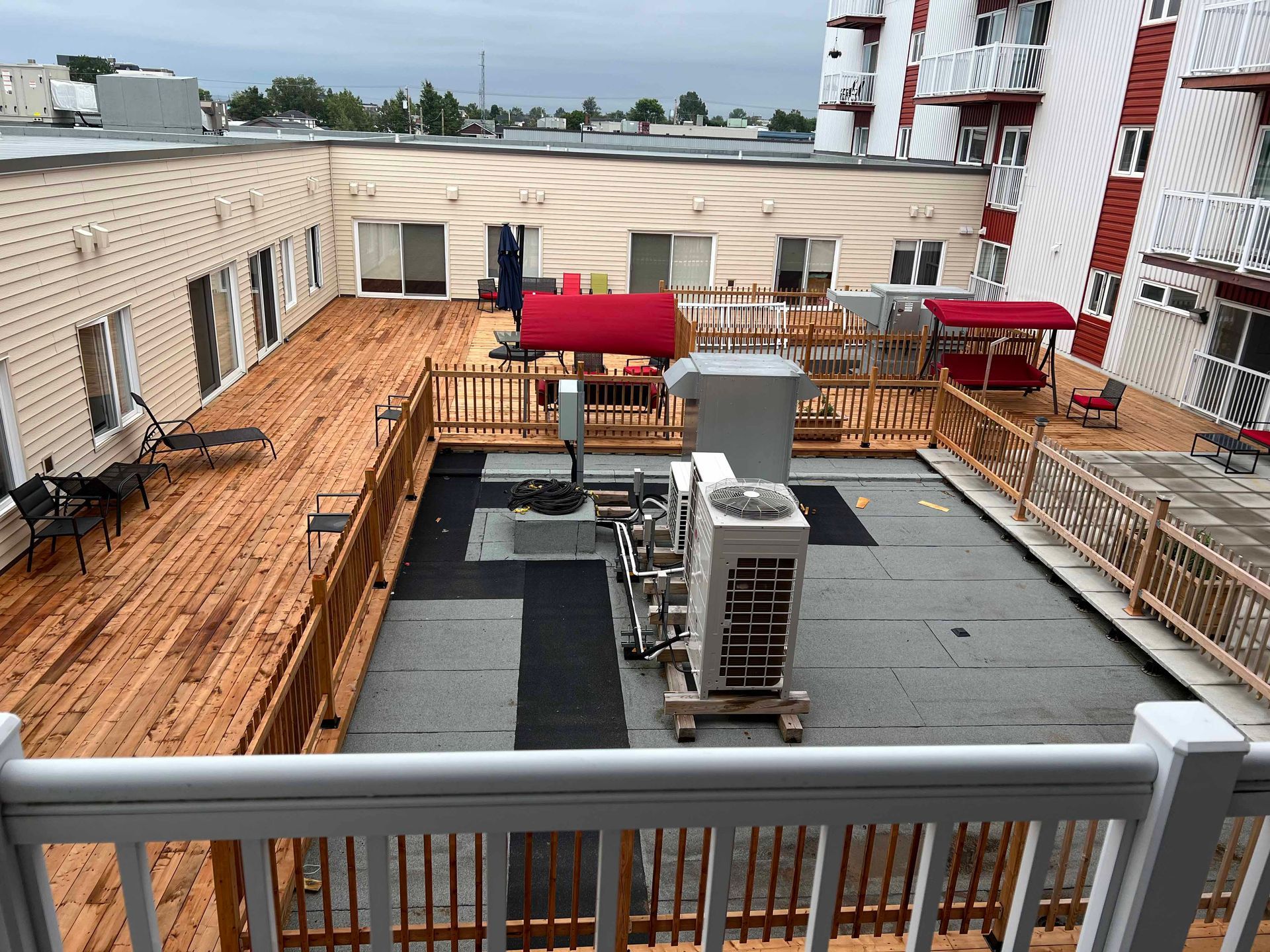 Toit-terrasse avec rambarde en bois et équipements techniques. On aperçoit des auvents rouges et un bâtiment à façade blanche et rouge.