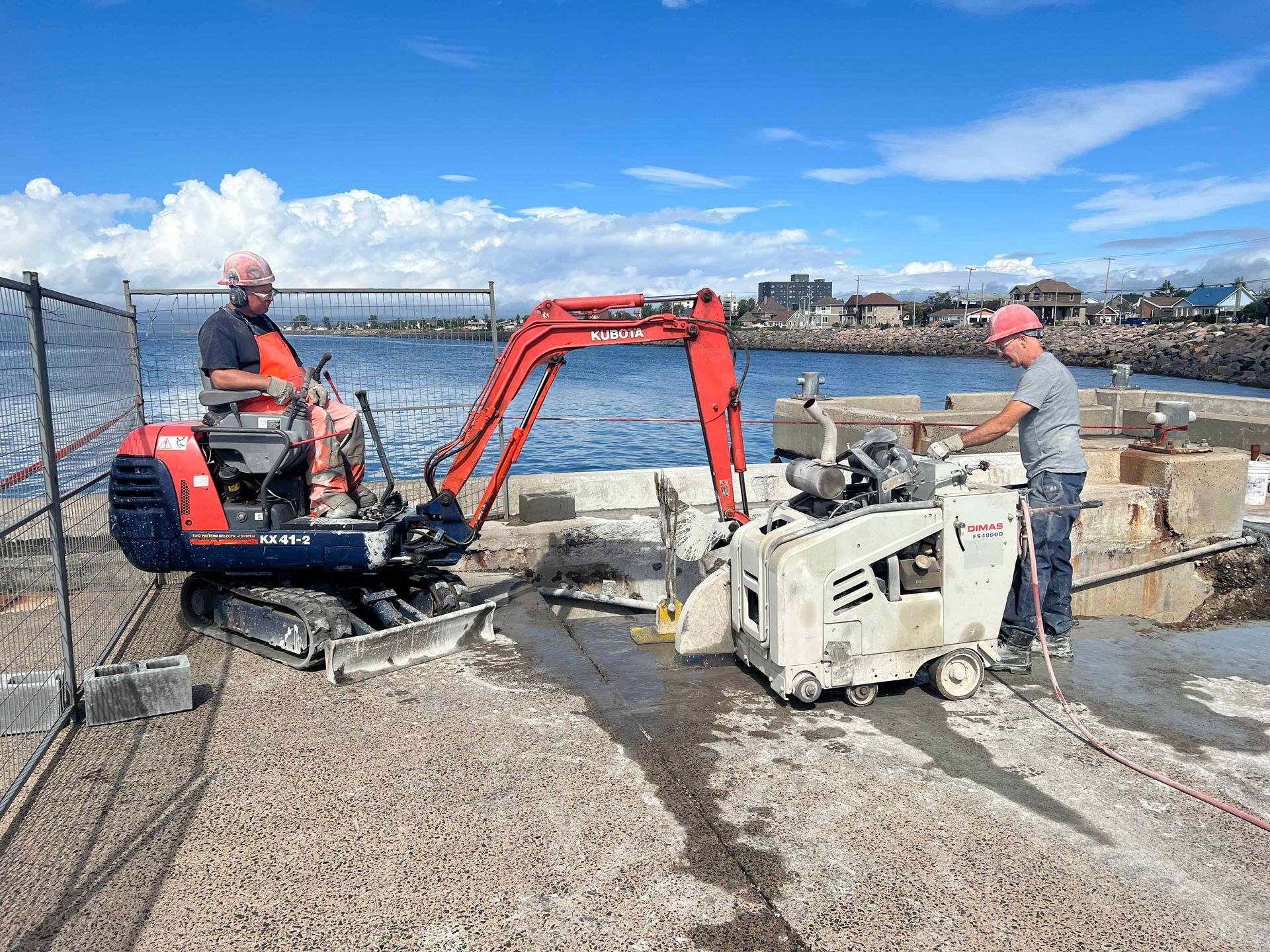 Deux ouvriers utilisent des engins de chantier sur un quai en béton près de l'eau : une mini-pelle et une scie à béton.
