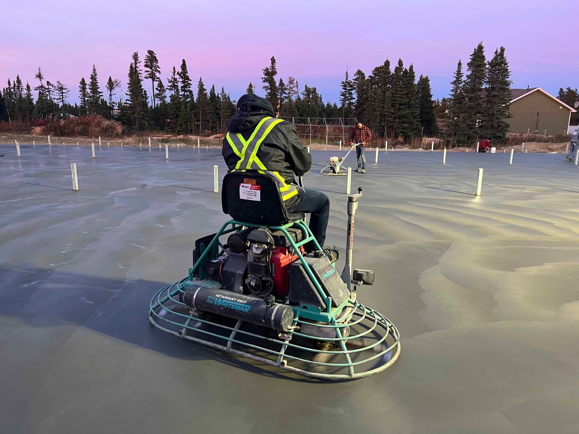 Une personne maniant une truelle mécanique à béton lisse une grande surface de béton fraîchement coulée à l'extérieur, au crépuscule.
