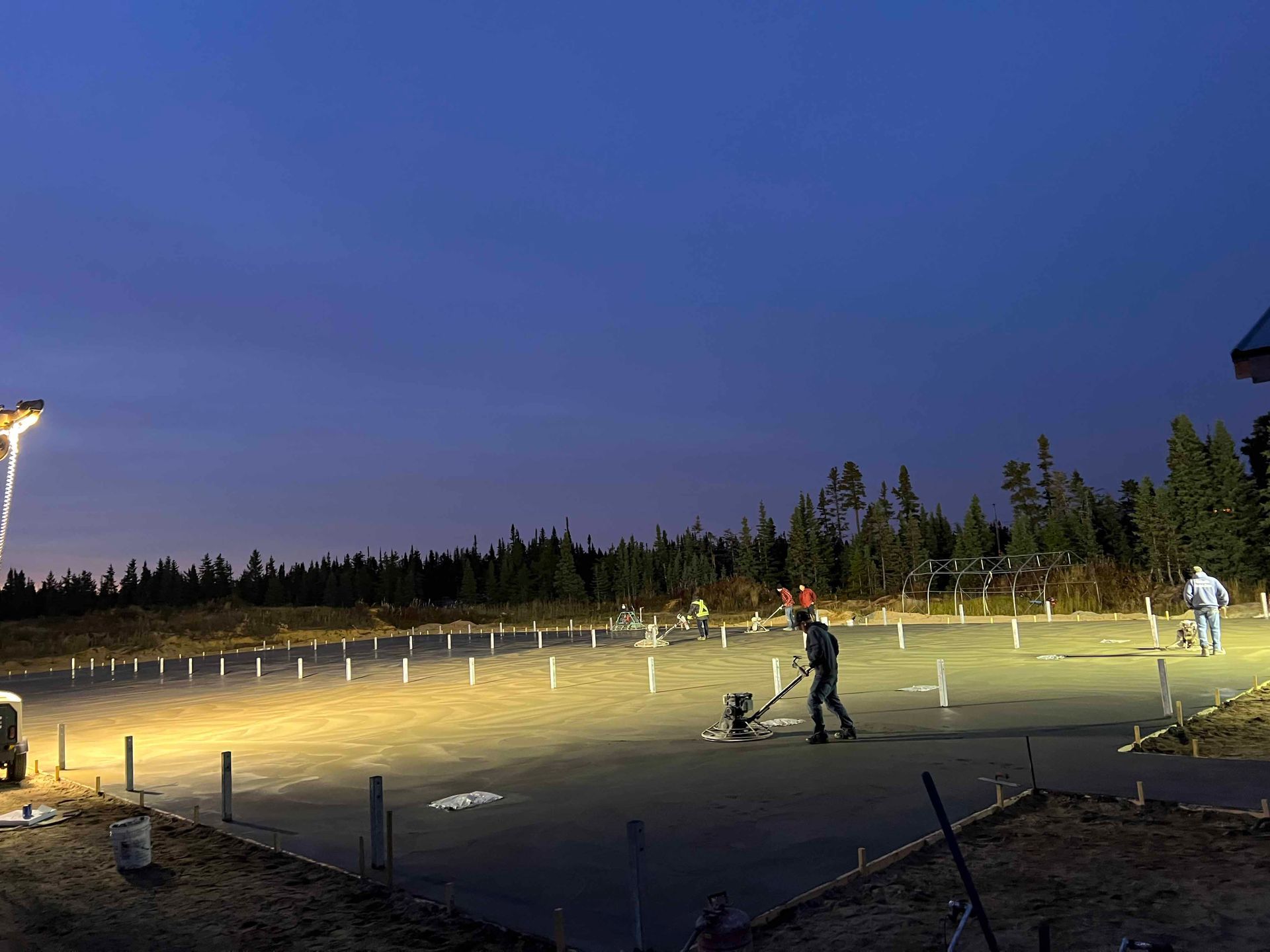 Des ouvriers du bâtiment lissent du béton au crépuscule, éclairés par des projecteurs, avec une forêt en arrière-plan.