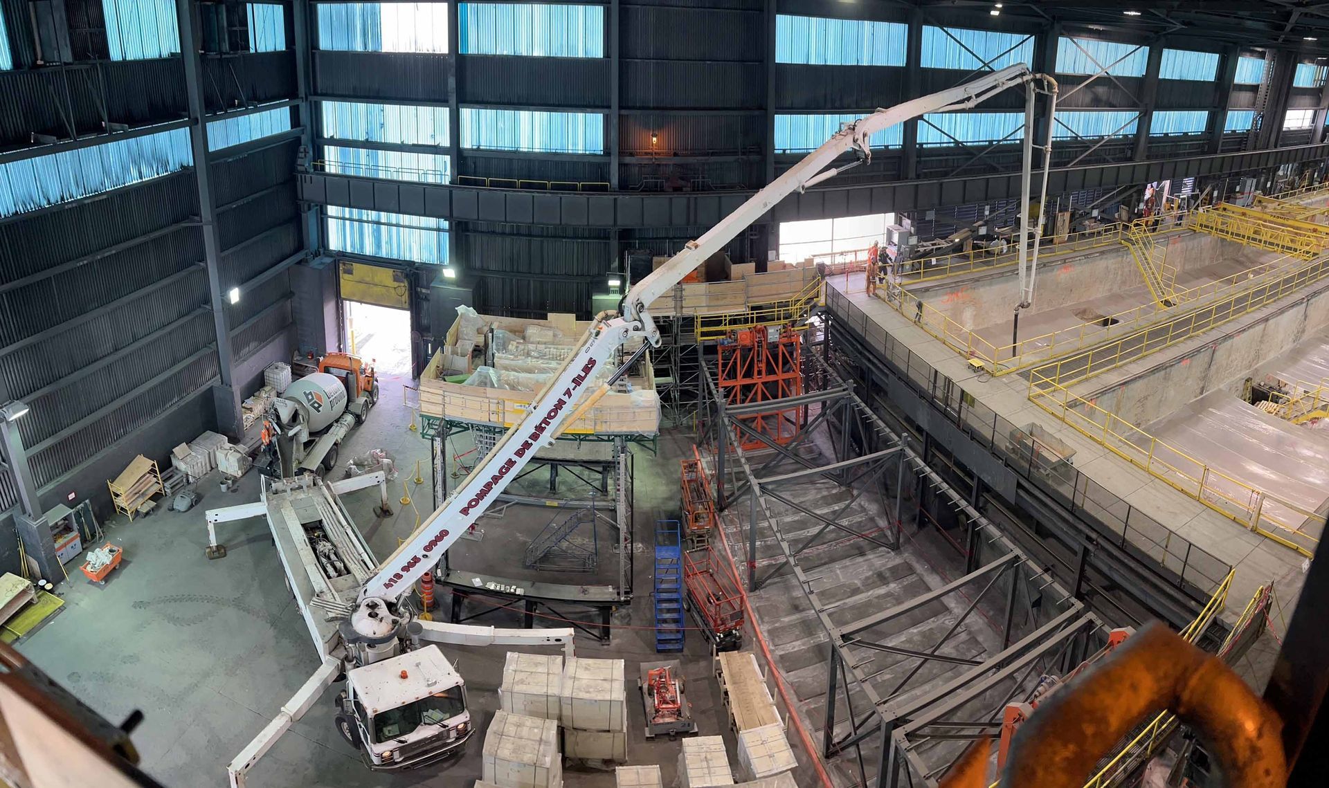 Des ouvriers du bâtiment utilisent une grue dans un entrepôt pour déplacer du matériel, près d'une grande structure en béton.