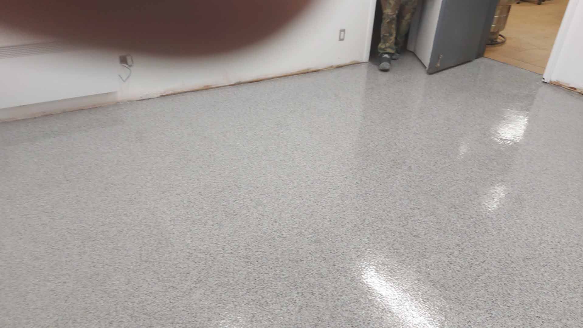 Un sol en époxy gris dans une pièce, avec un mur blanc et des pieds visibles en arrière-plan.