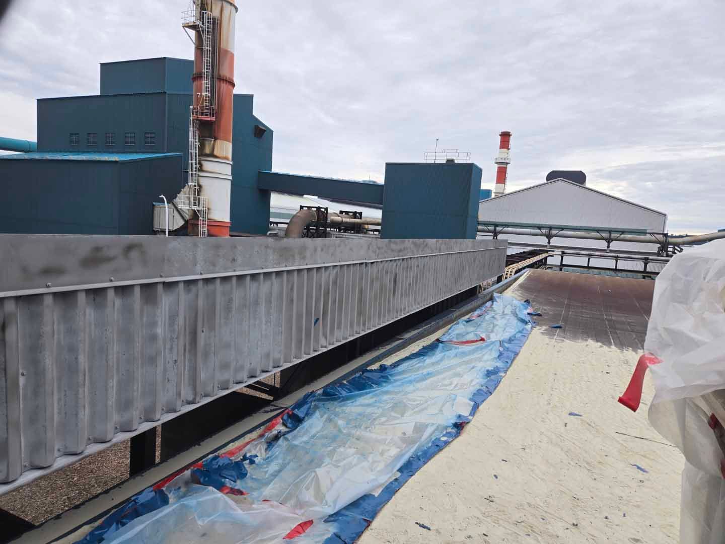 Toit d'un bâtiment industriel avec une grande structure métallique, des bâches bleues et des cheminées d'usine se détachant sur un ciel nuageux.