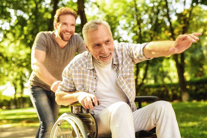 A pensioner on a wheelchair and his adult son are walking around the park.