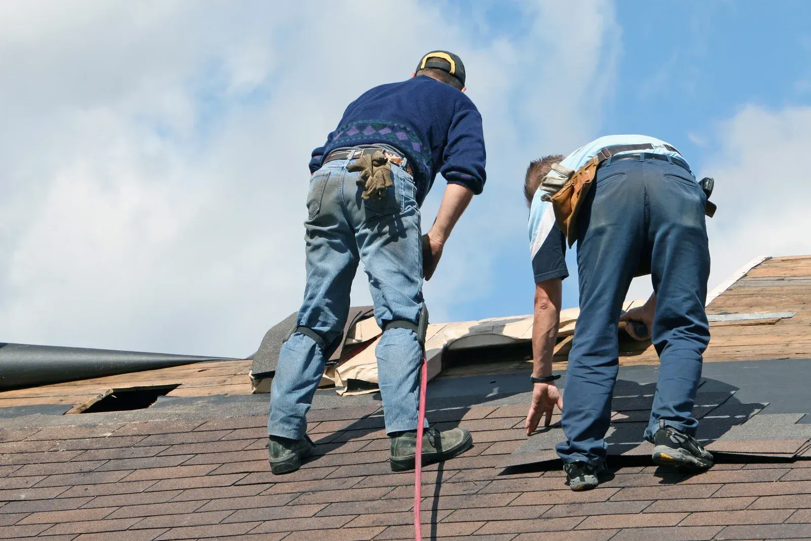 Two roofers repairing a roof, wearing work belts, under a partly cloudy sky.