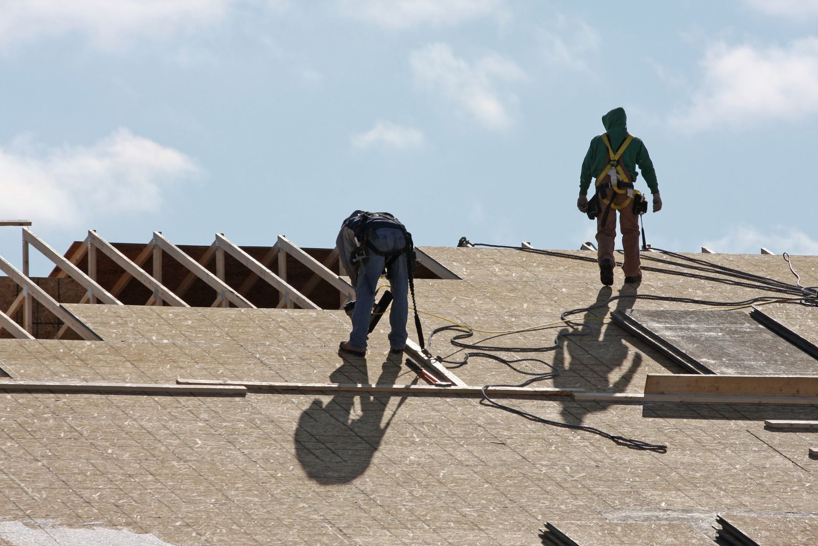 Two roofers working on a roof under a blue sky, one kneeling and one standing, both wearing safety harnesses.