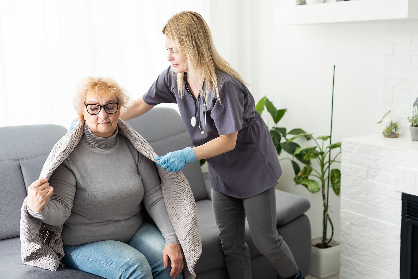 A nurse is helping an elderly woman sitting on a couch with a blanket.