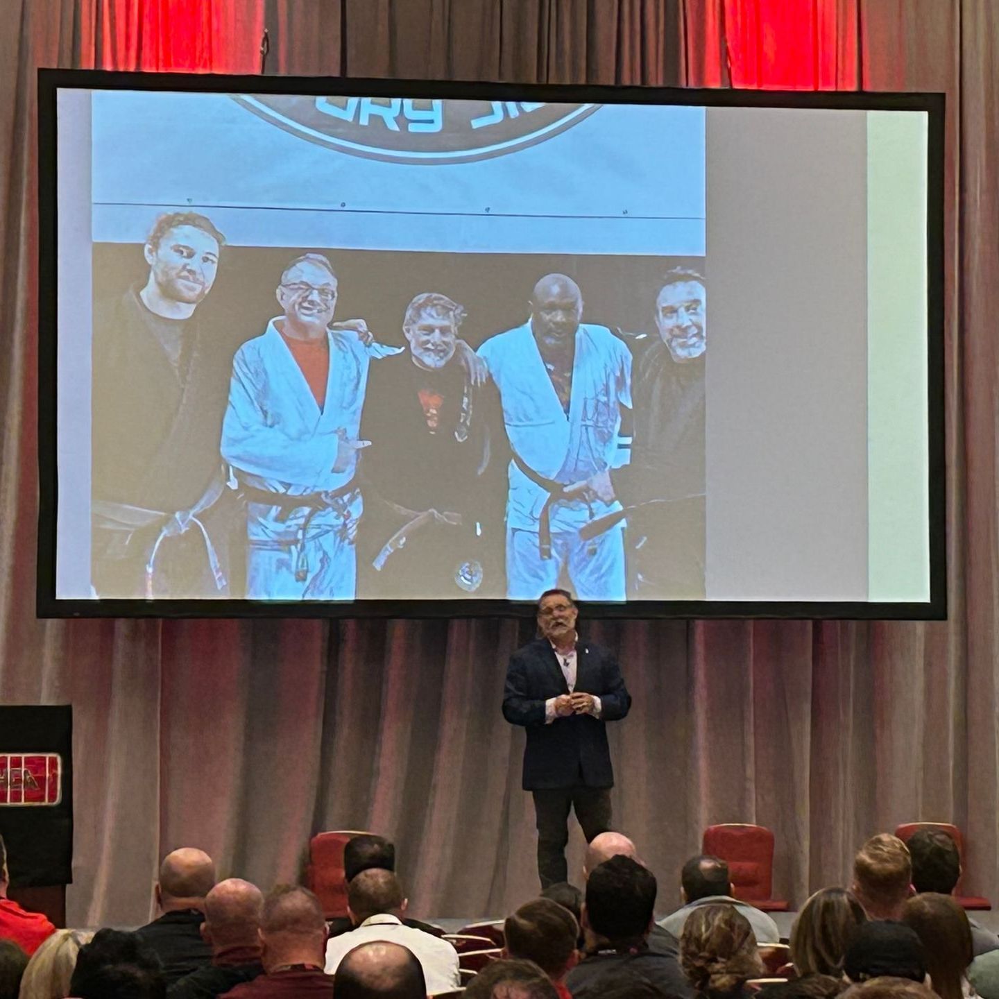 A man is giving a presentation in front of a large screen with the word karate on it
