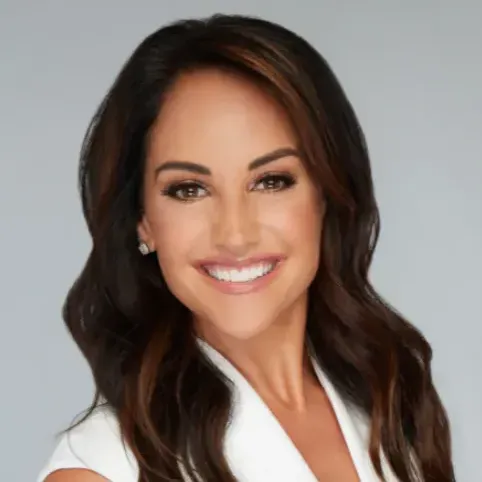 A woman with long brown hair is wearing a white jacket and smiling.