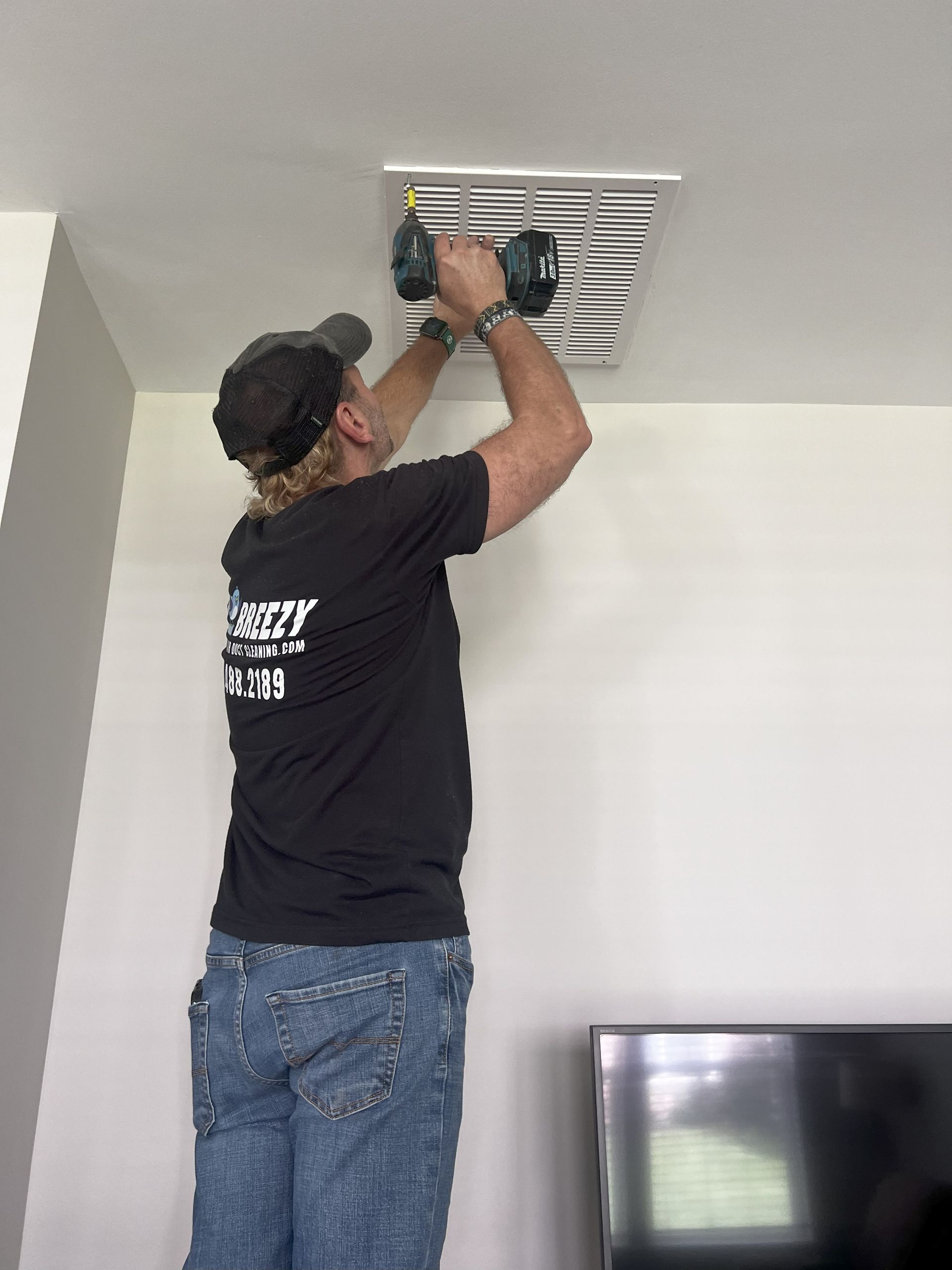Man in a black shirt uses a drill to install a ceiling vent. He stands on a platform.