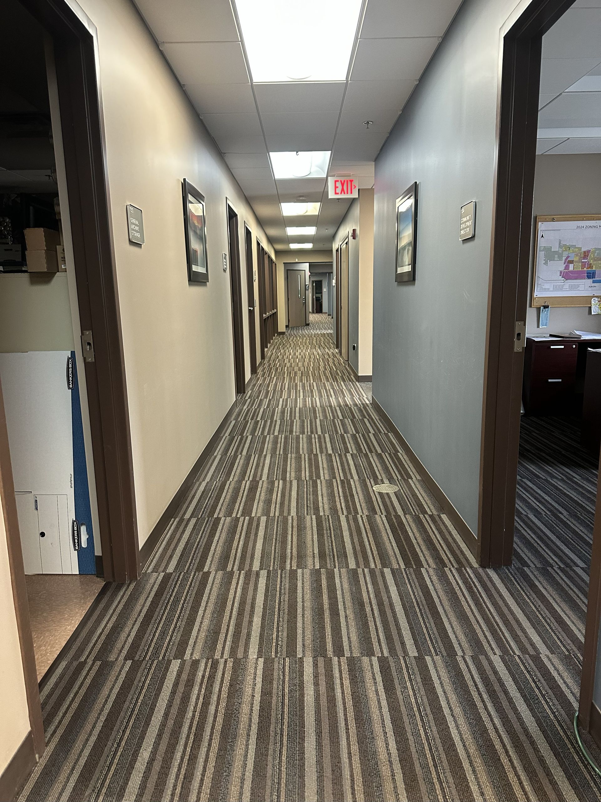 Long office hallway with doors on both sides, neutral-toned walls, patterned carpet, and overhead lighting.