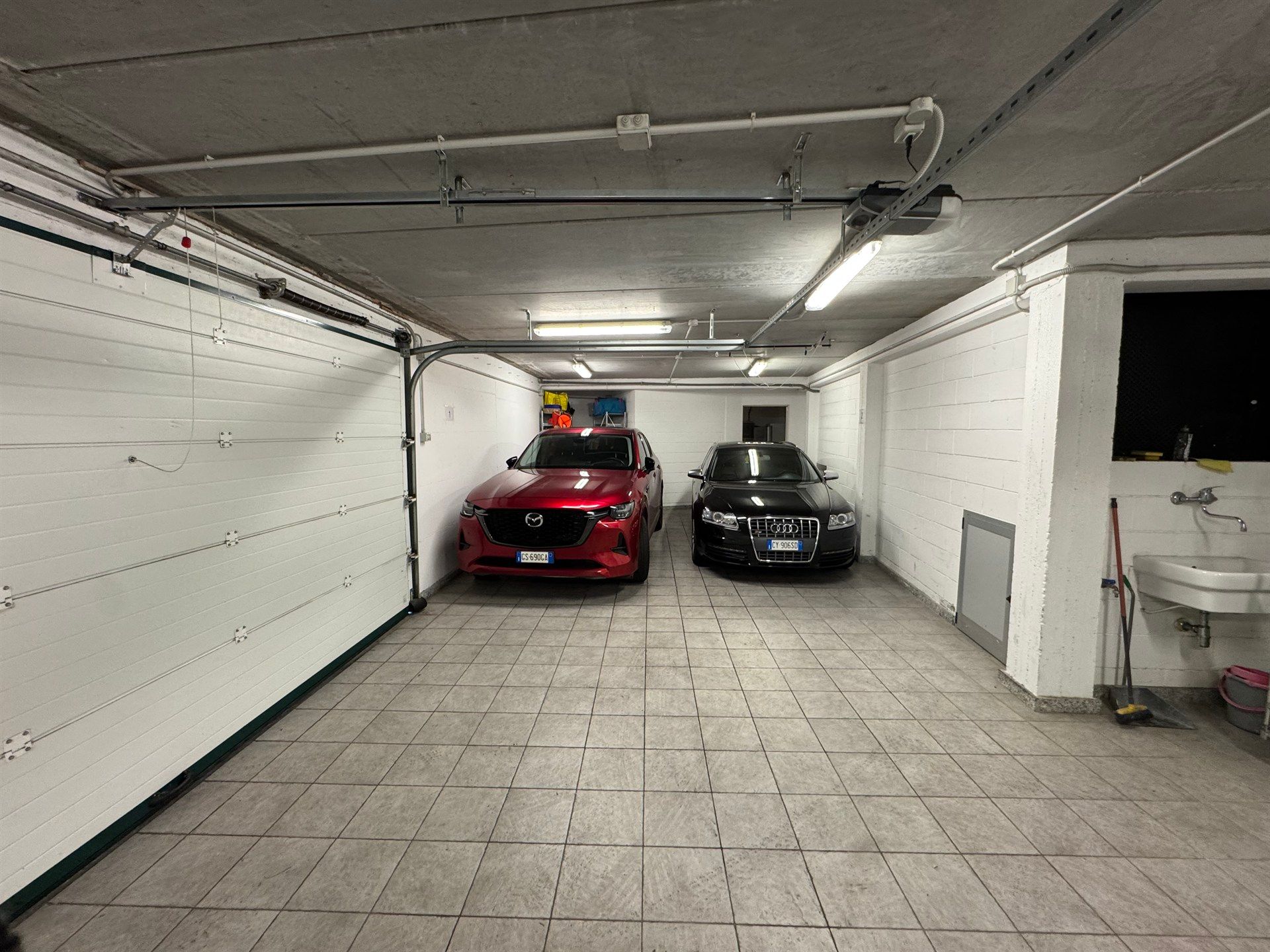 Two cars are parked in a garage with a sink.