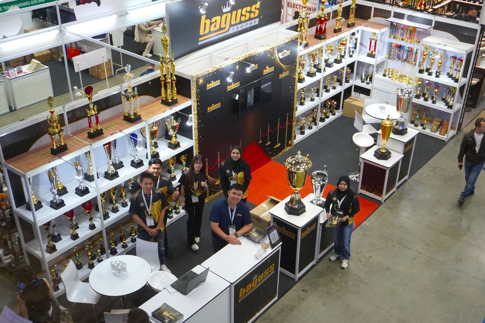 Group photo of Baguss Enterprise team at exhibition booth during Malaysia Gifts Fair 2025, featuring custom trophies and medals