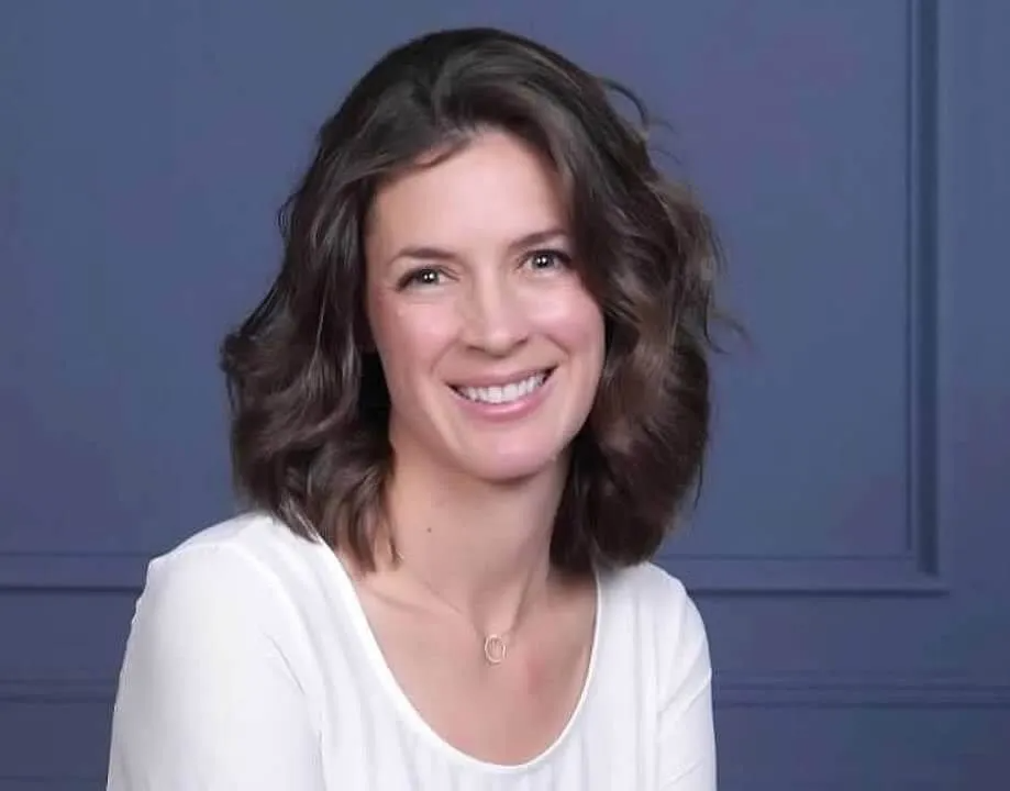 Woman with brown wavy hair smiles in front of a blue background, wearing a white top and necklace.