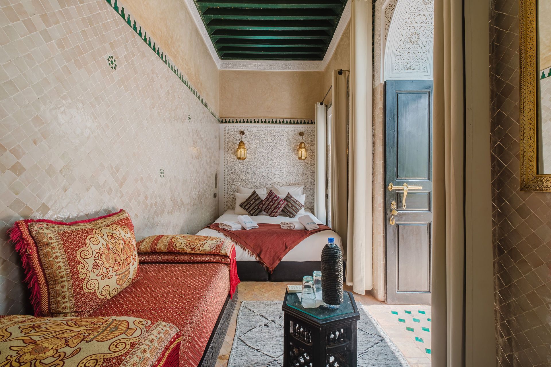 Cozy Moroccan bedroom: red couch, bed, small table with drinks, tiled walls, doorway to balcony.