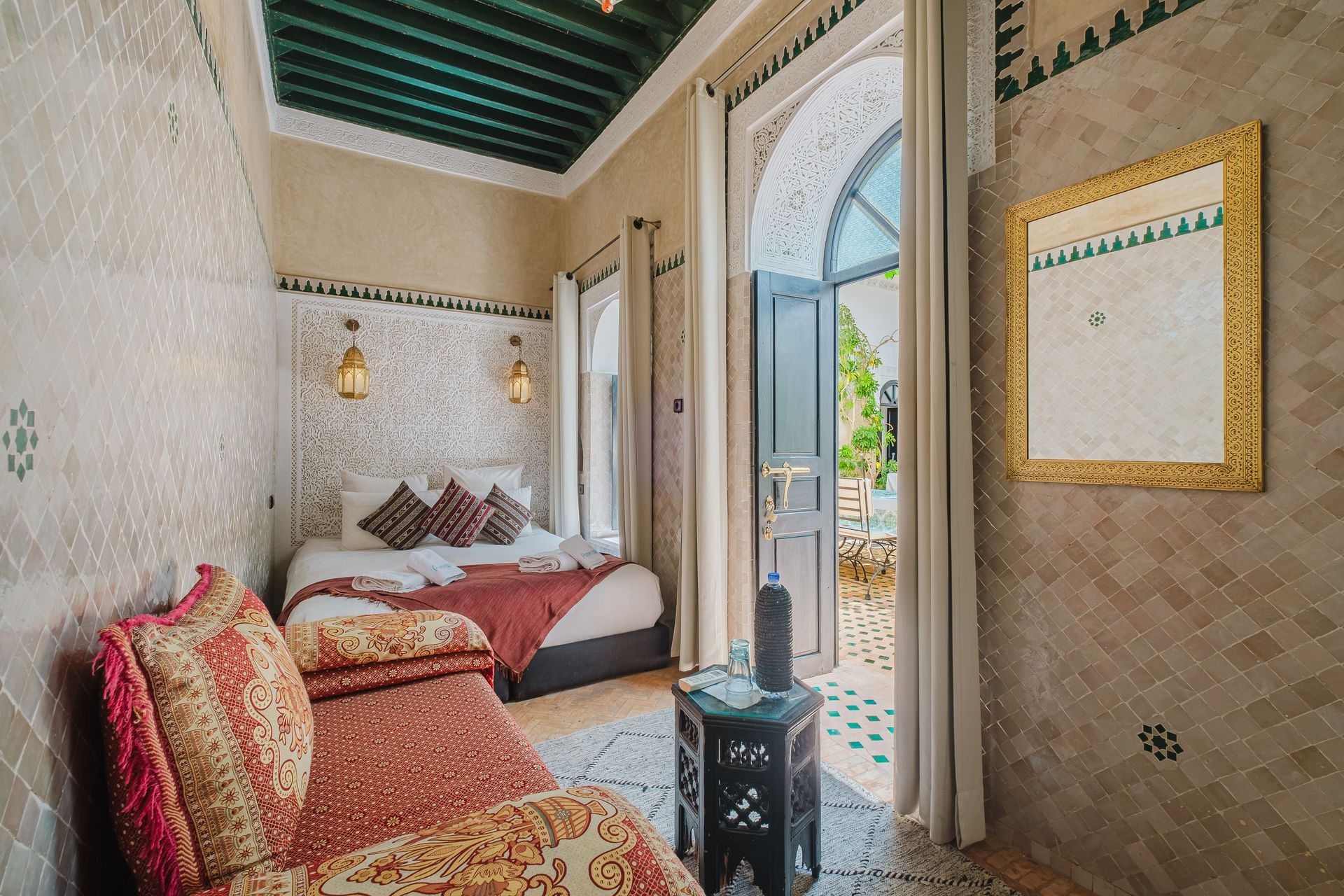 Moroccan-style bedroom with a bed, seating, doorway to patio. Beige tile walls, gold mirror, green ceiling.