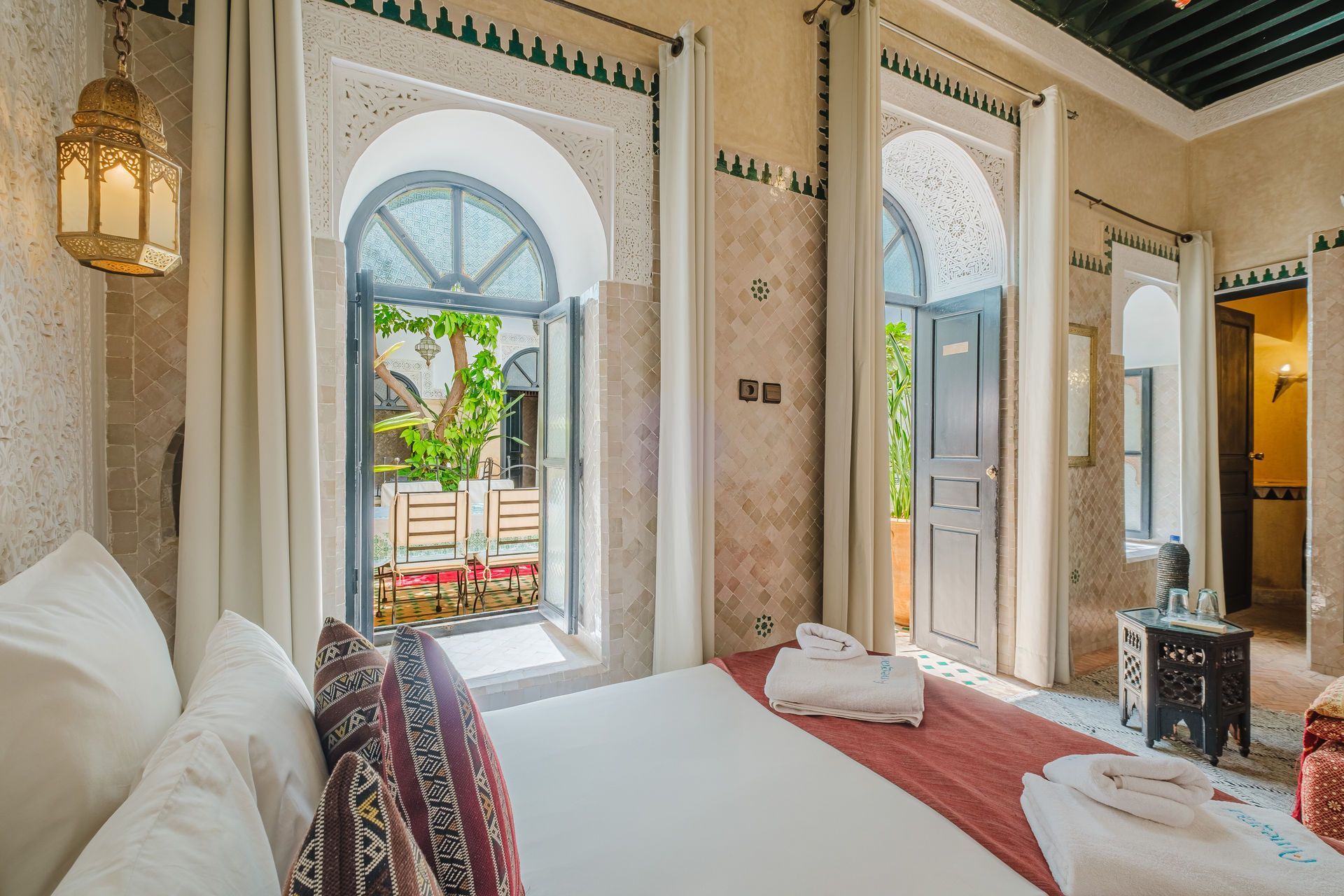 Bedroom with arched doors opening to a courtyard. Bed has pillows and a red runner. Lantern hangs to the left.