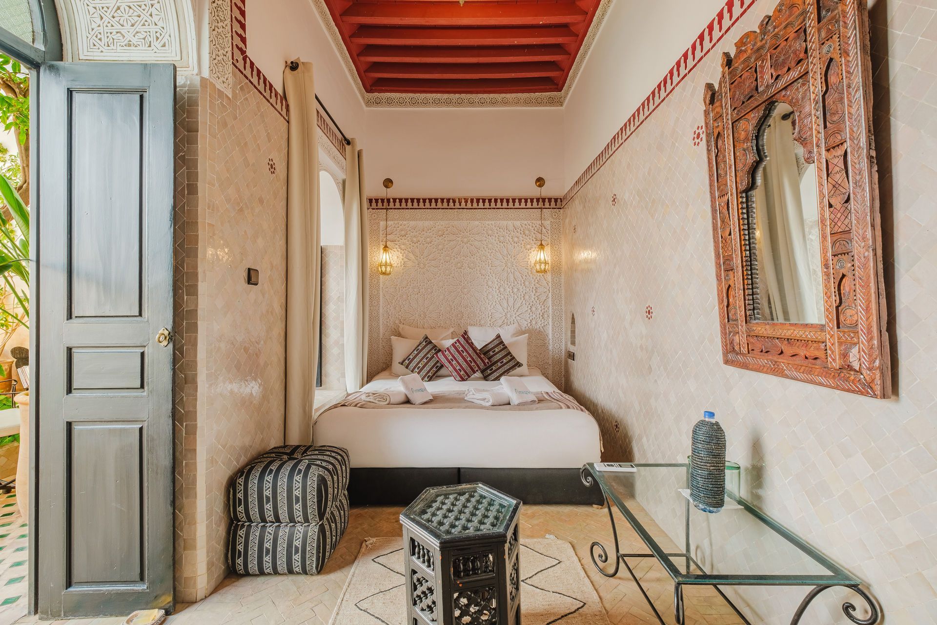 Bedroom in traditional Moroccan style with bed, patterned rug, carved wood mirror, and open doorway.