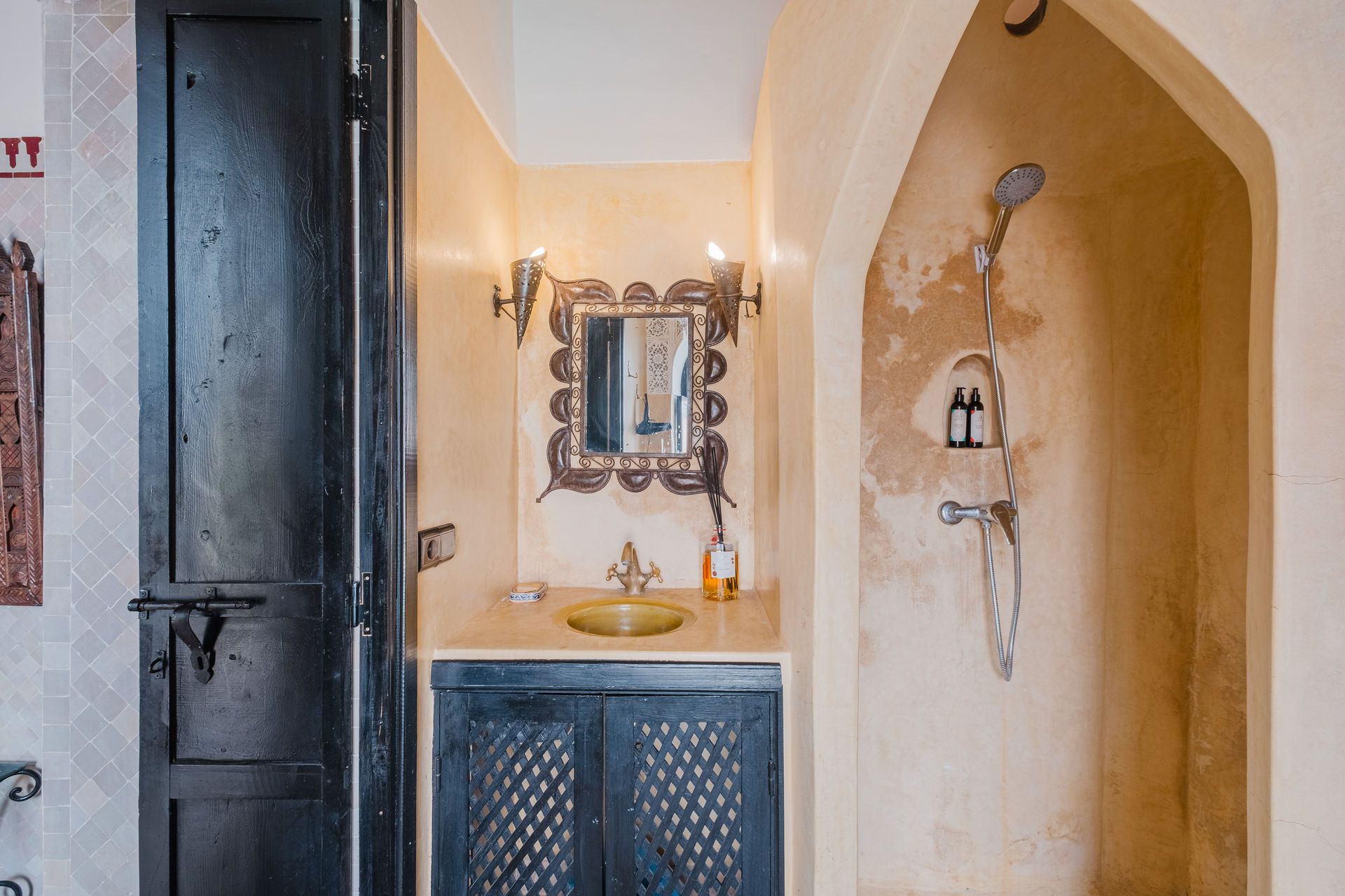 Moroccan-style bathroom: black door, shower alcove, ornate mirror, sink, and cabinet, all in muted tones.