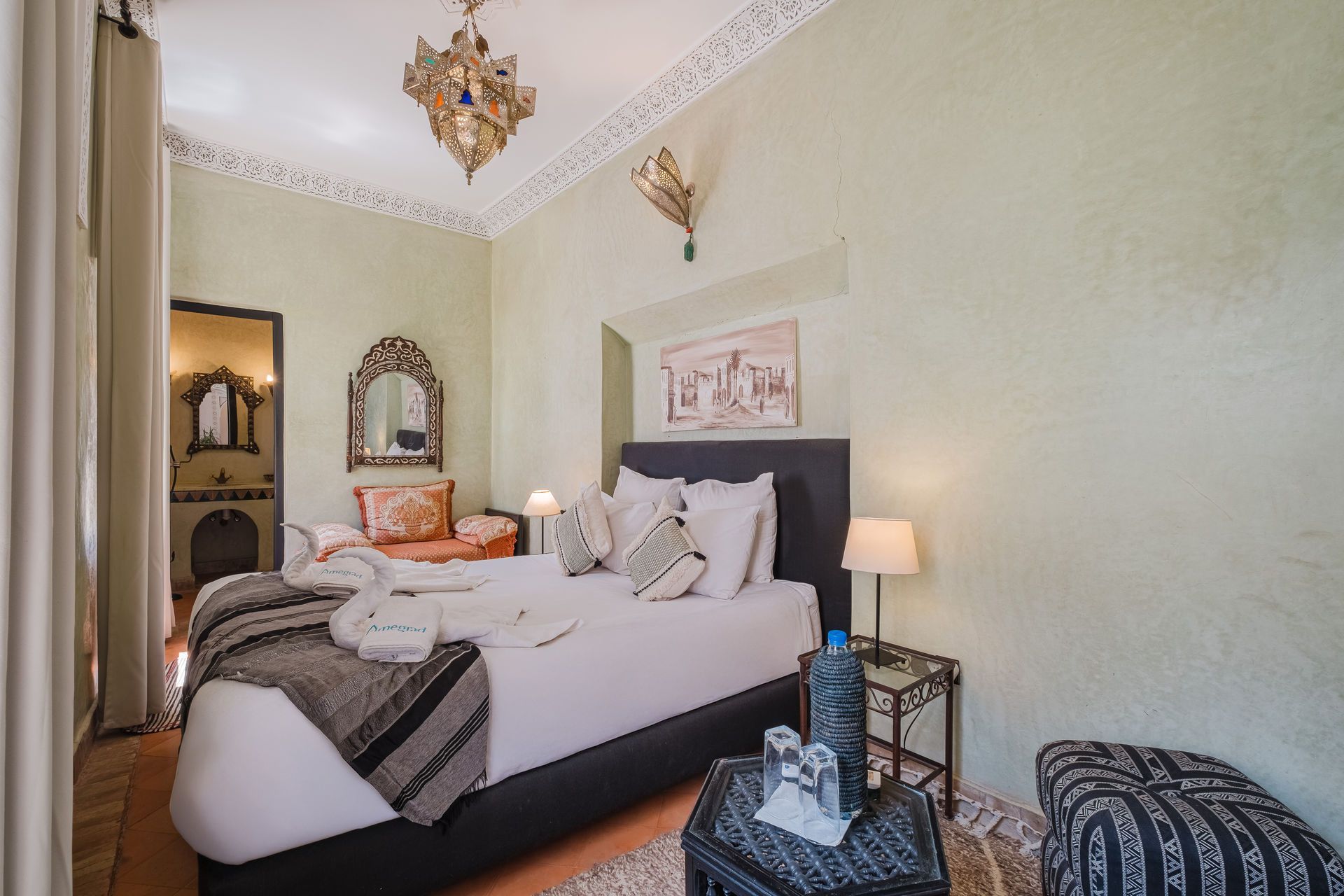 Bedroom with a large bed, ornate lighting, light green walls, and a seating area with a decorative mirror.