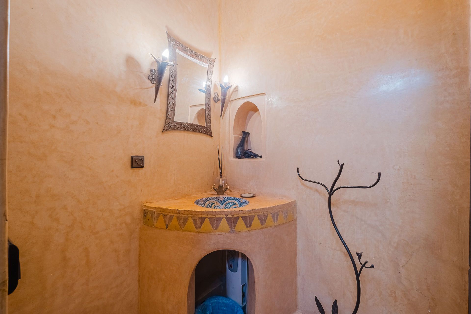 Tan-walled bathroom with arched sink, mosaic tile countertop, and ornate mirror; decorative metal branch.