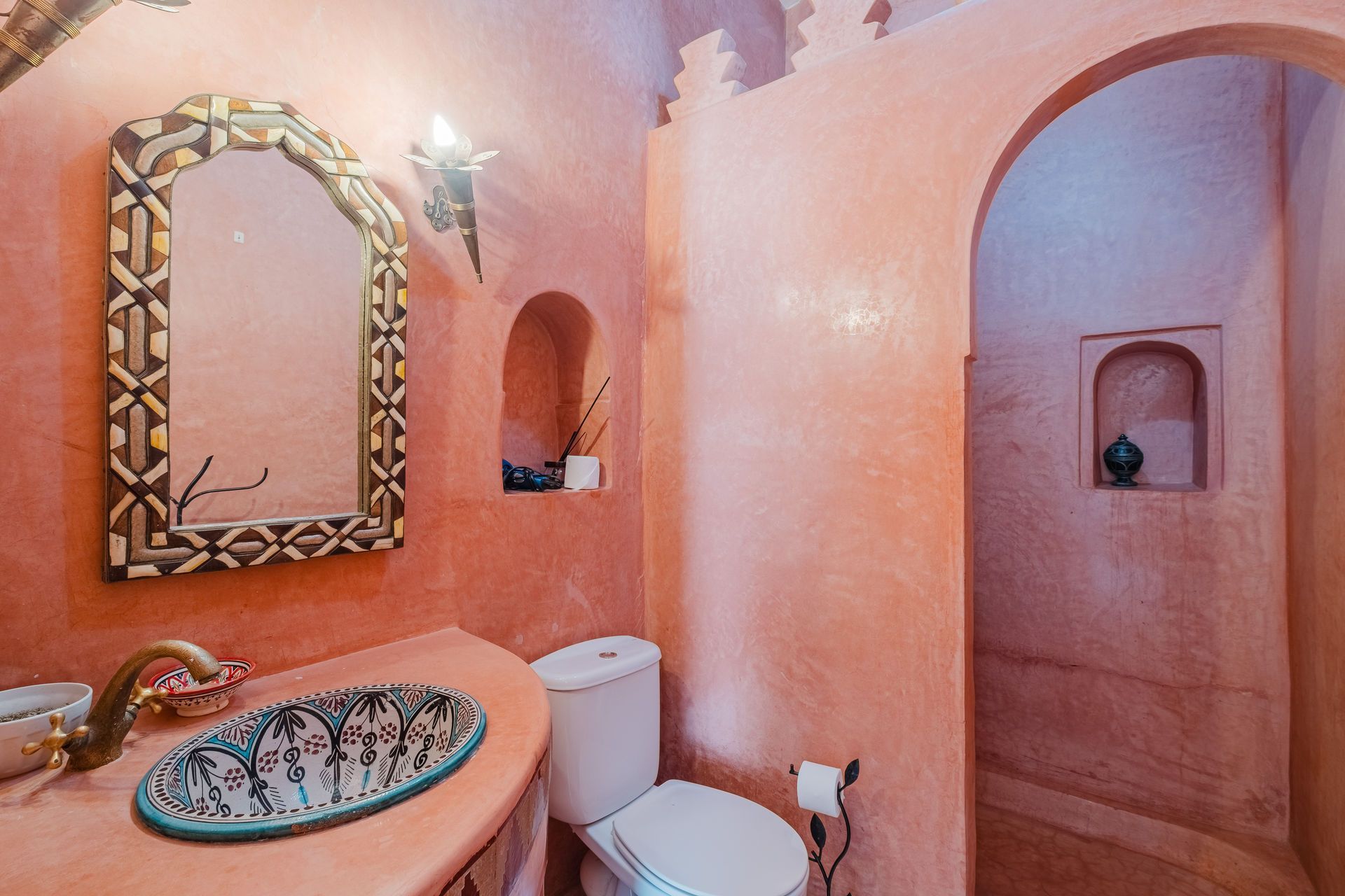 Pink-walled bathroom with ornate mirror and sink; arched doorway leads to a shower.
