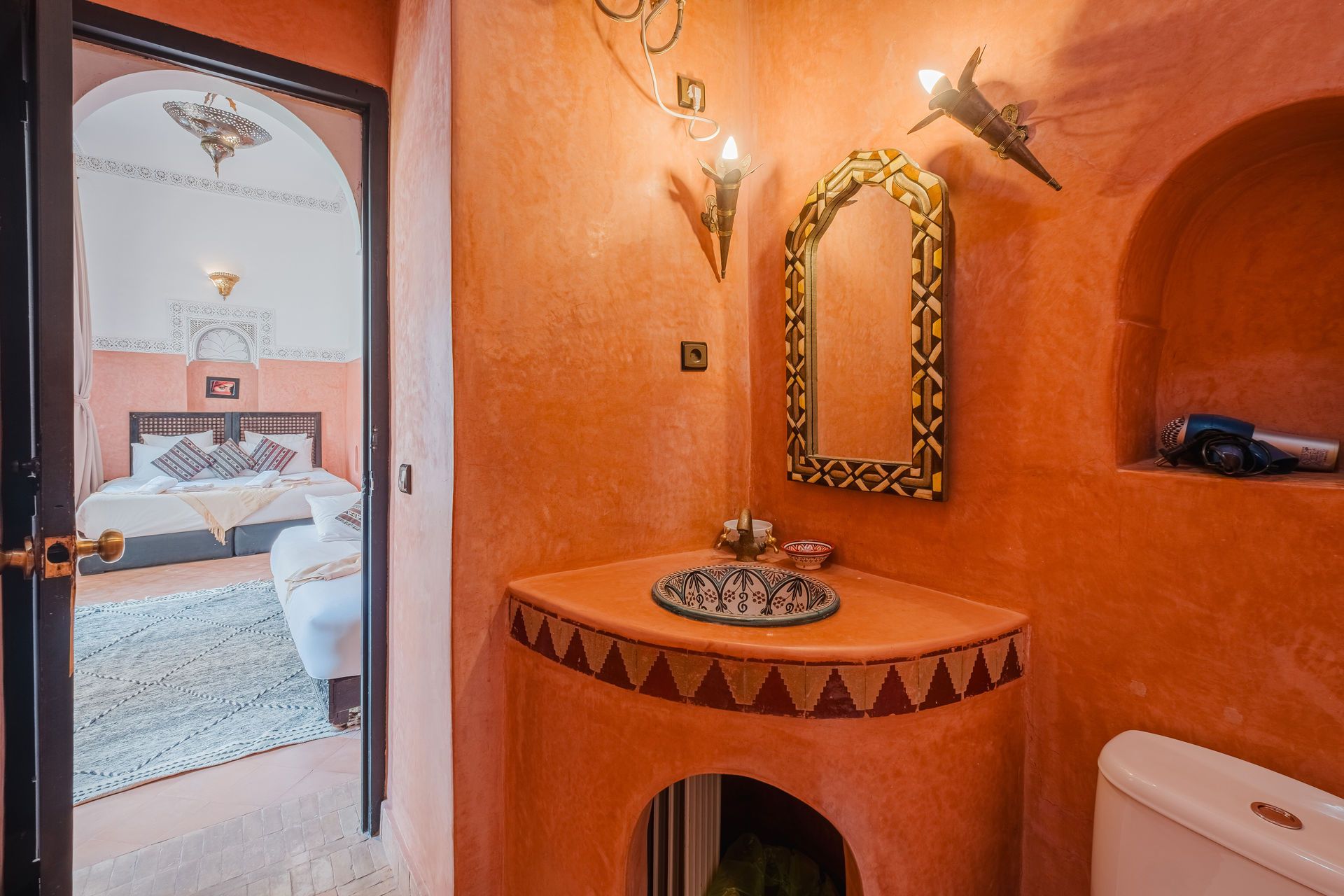 A Moroccan-style bathroom with orange walls, arched doorway, and a view of the bedroom.
