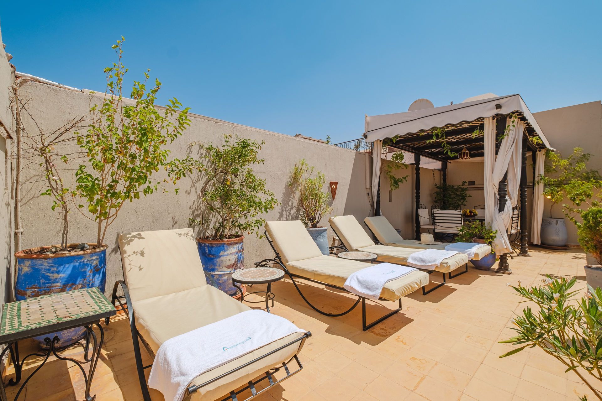 Rooftop patio with lounge chairs, shaded pergola, and potted plants under a bright blue sky.