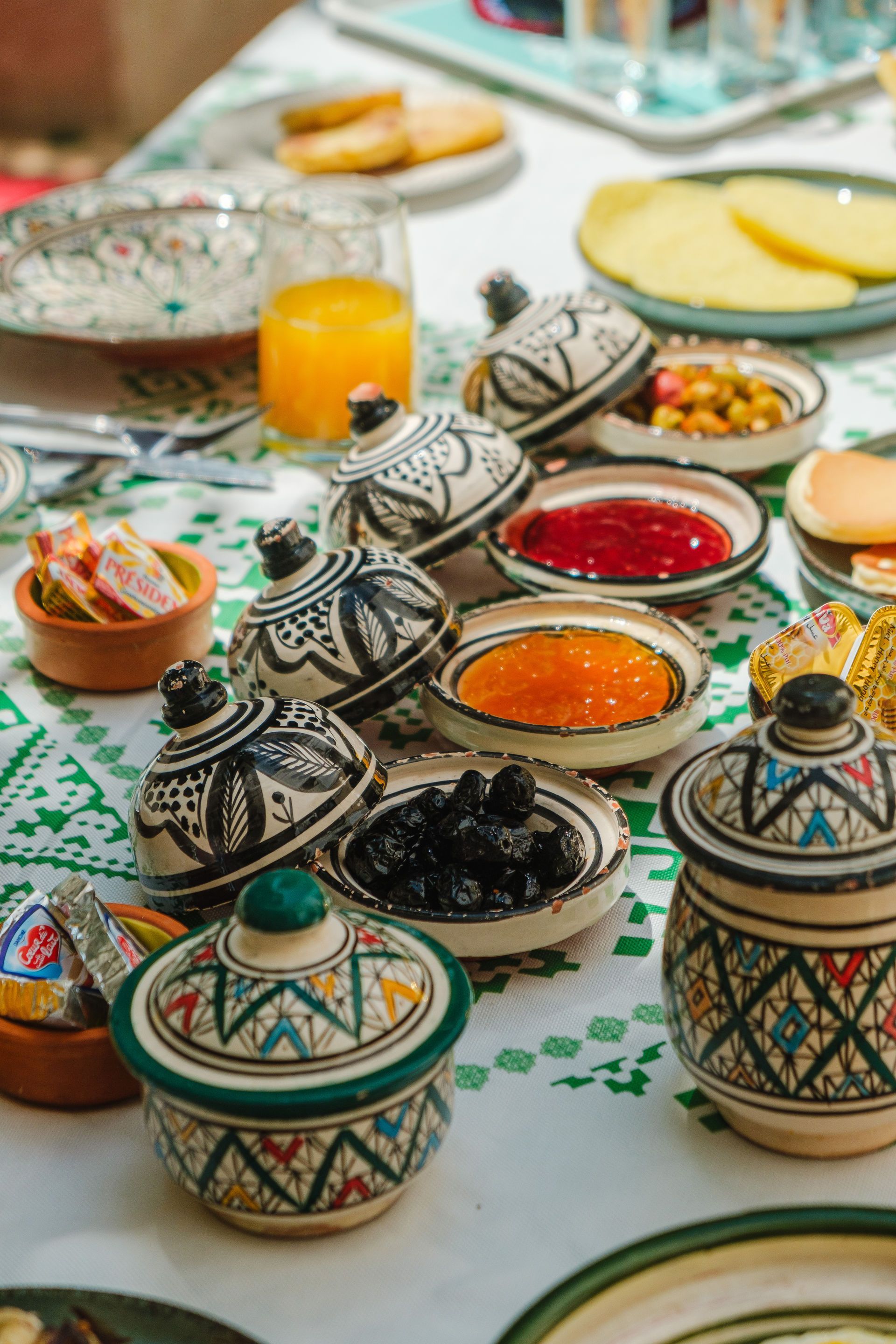 Breakfast table with decorative bowls of jams, olives, and breads on a white tablecloth with a green floral pattern.