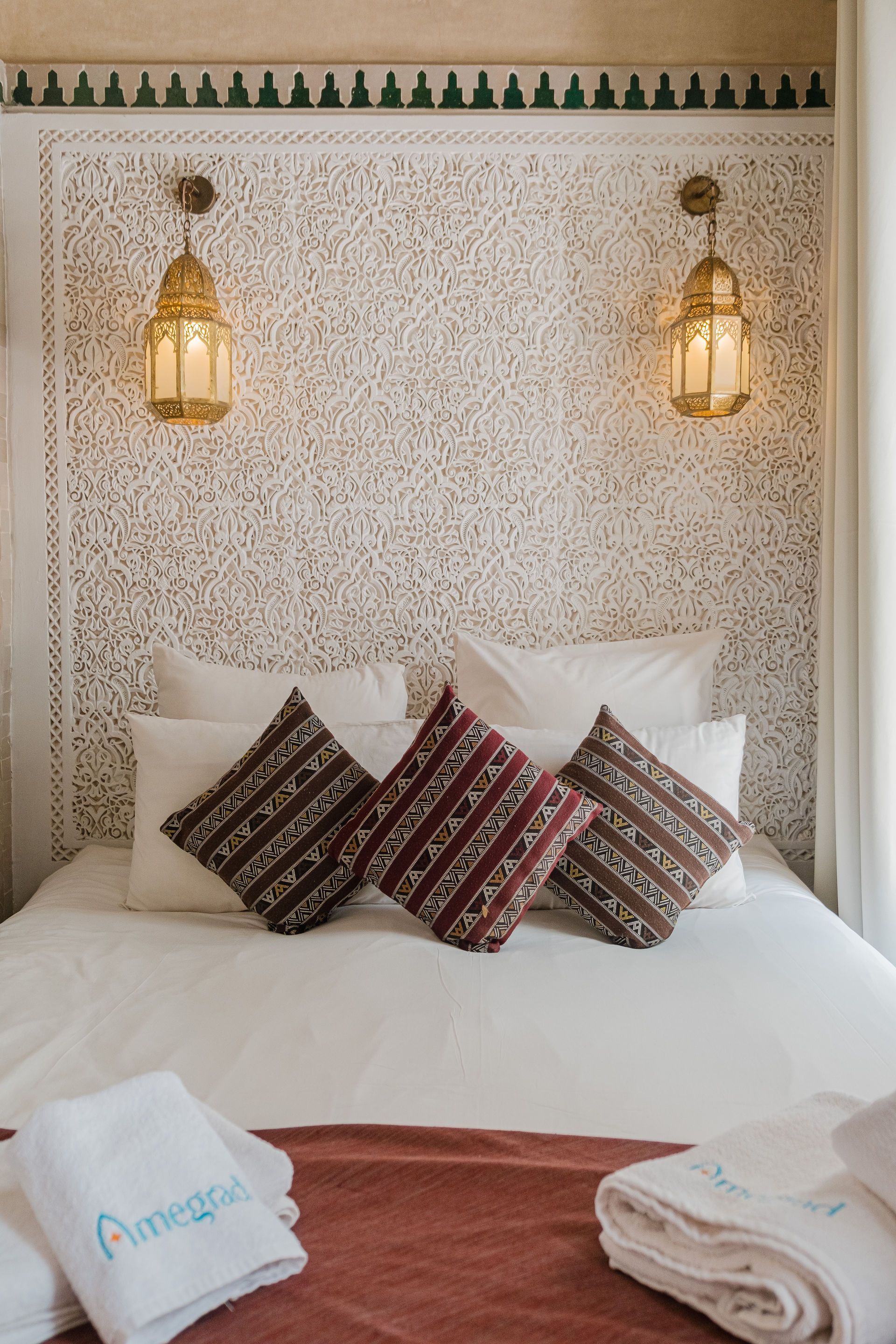 Bed with white linens and patterned throw pillows, against a carved white headboard with hanging lanterns.