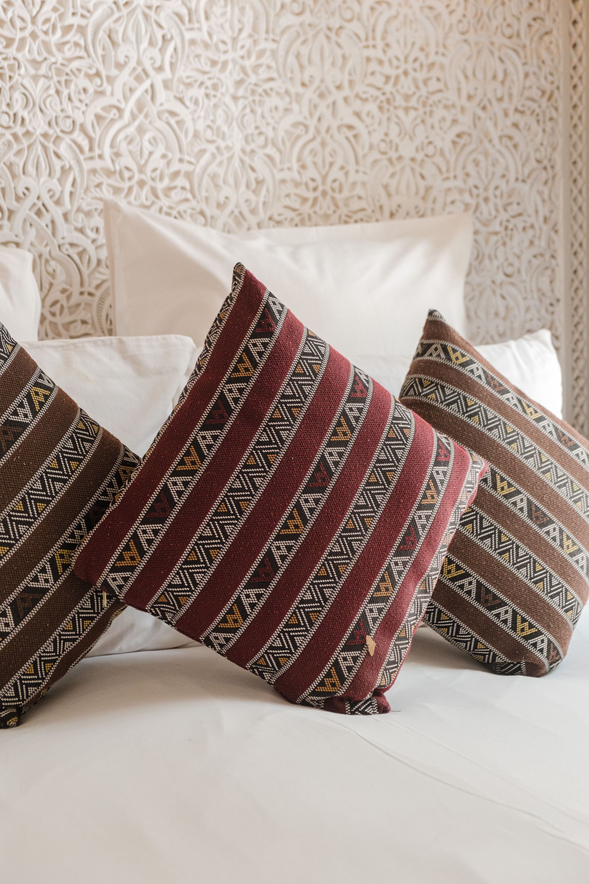 Three patterned throw pillows on a white bed, set against a carved white wall.